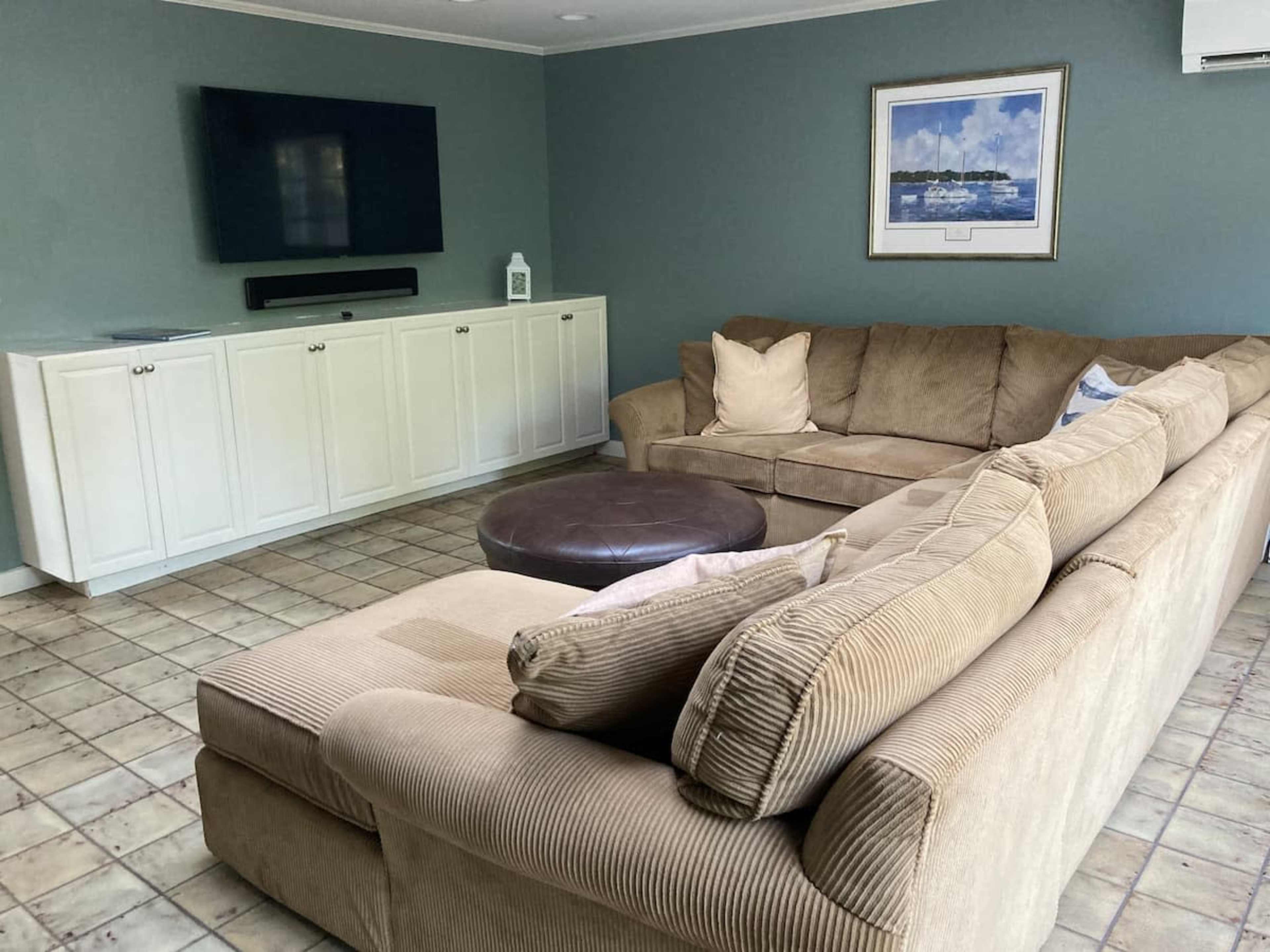 A spacious living room features a large sectional sofa, a round ottoman, a mounted TV, and light-colored cabinetry against a green wall.