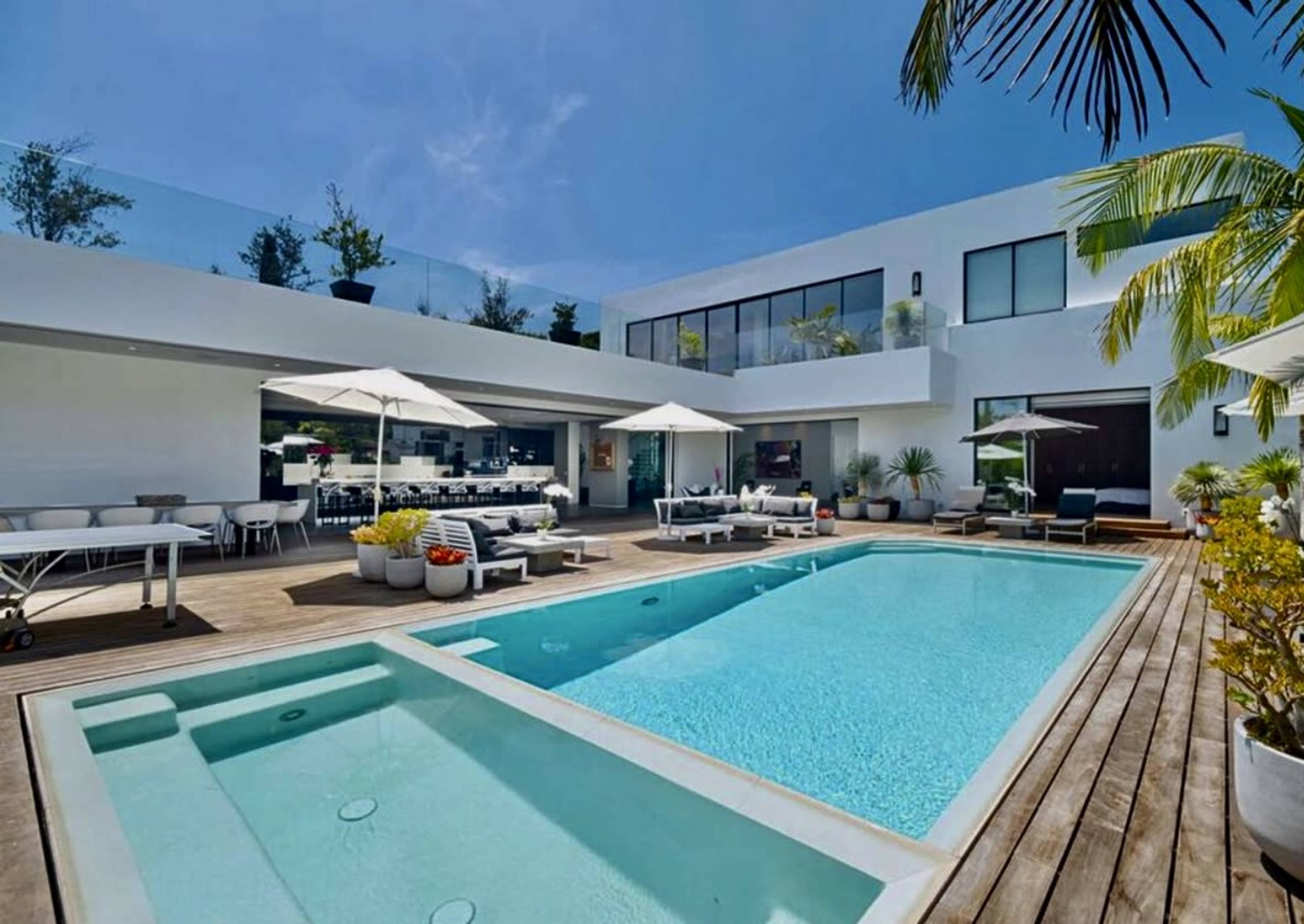 A modern home with a spacious patio featuring two swimming pools, lounge chairs, and outdoor dining areas surrounded by greenery.