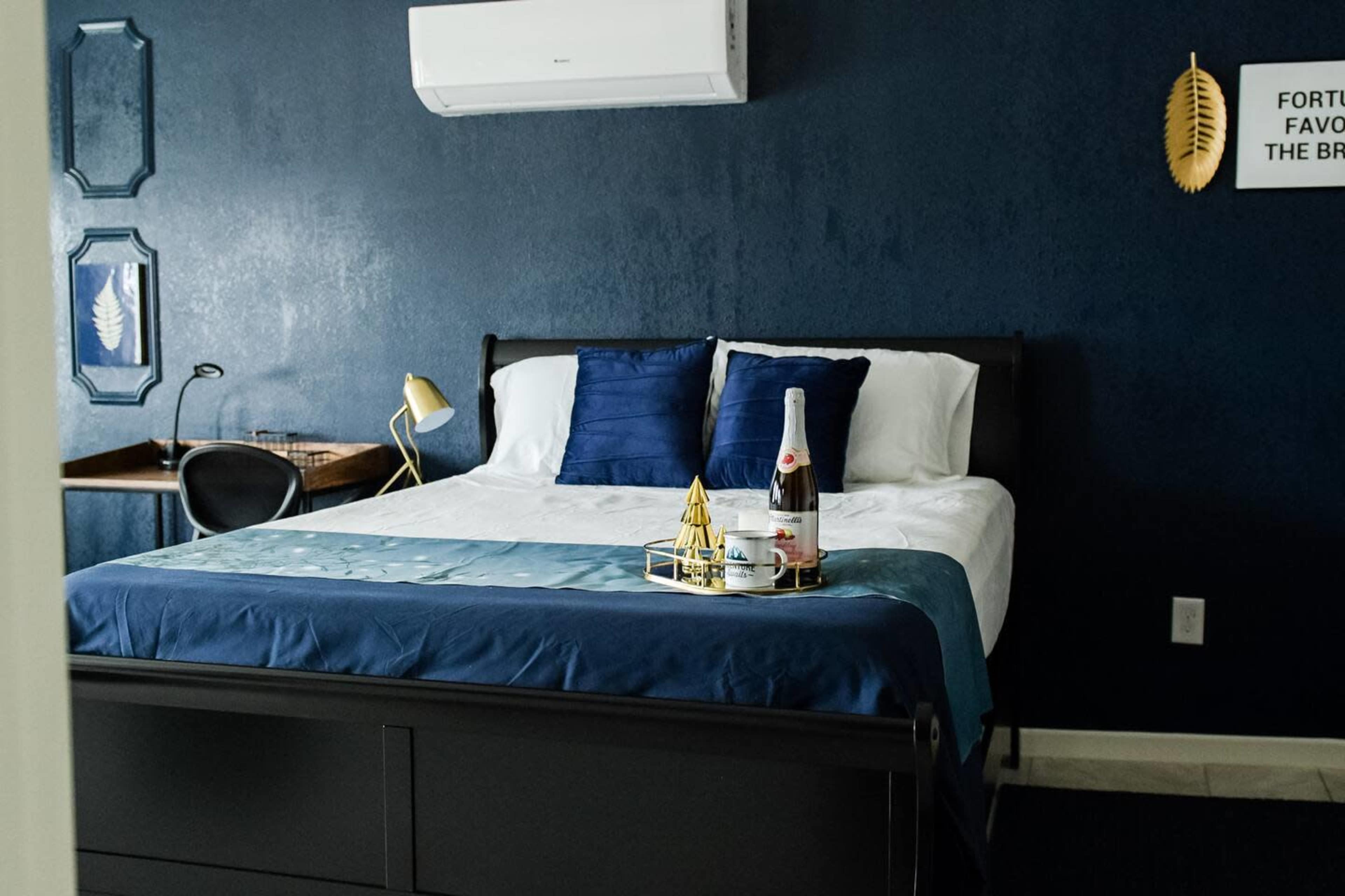 A neatly arranged bedroom features a dark blue wall, a queen-sized bed with white linens and blue accent pillows, a small desk, and a tray with a bottle of champagne and decorative items.