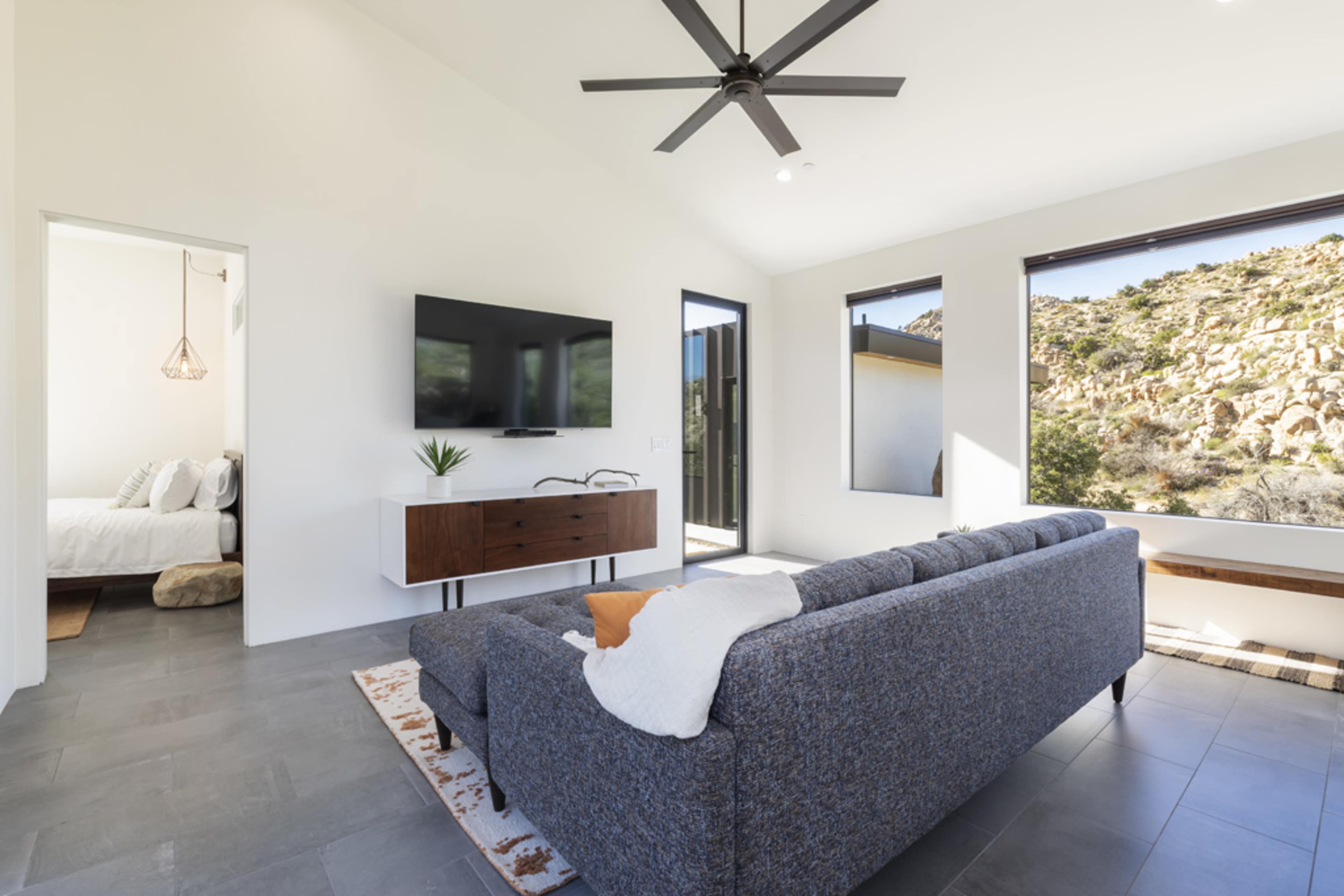 A modern living room features a gray sofa, a wooden TV stand with a flat-screen television, and large windows showcasing a rocky landscape outside.