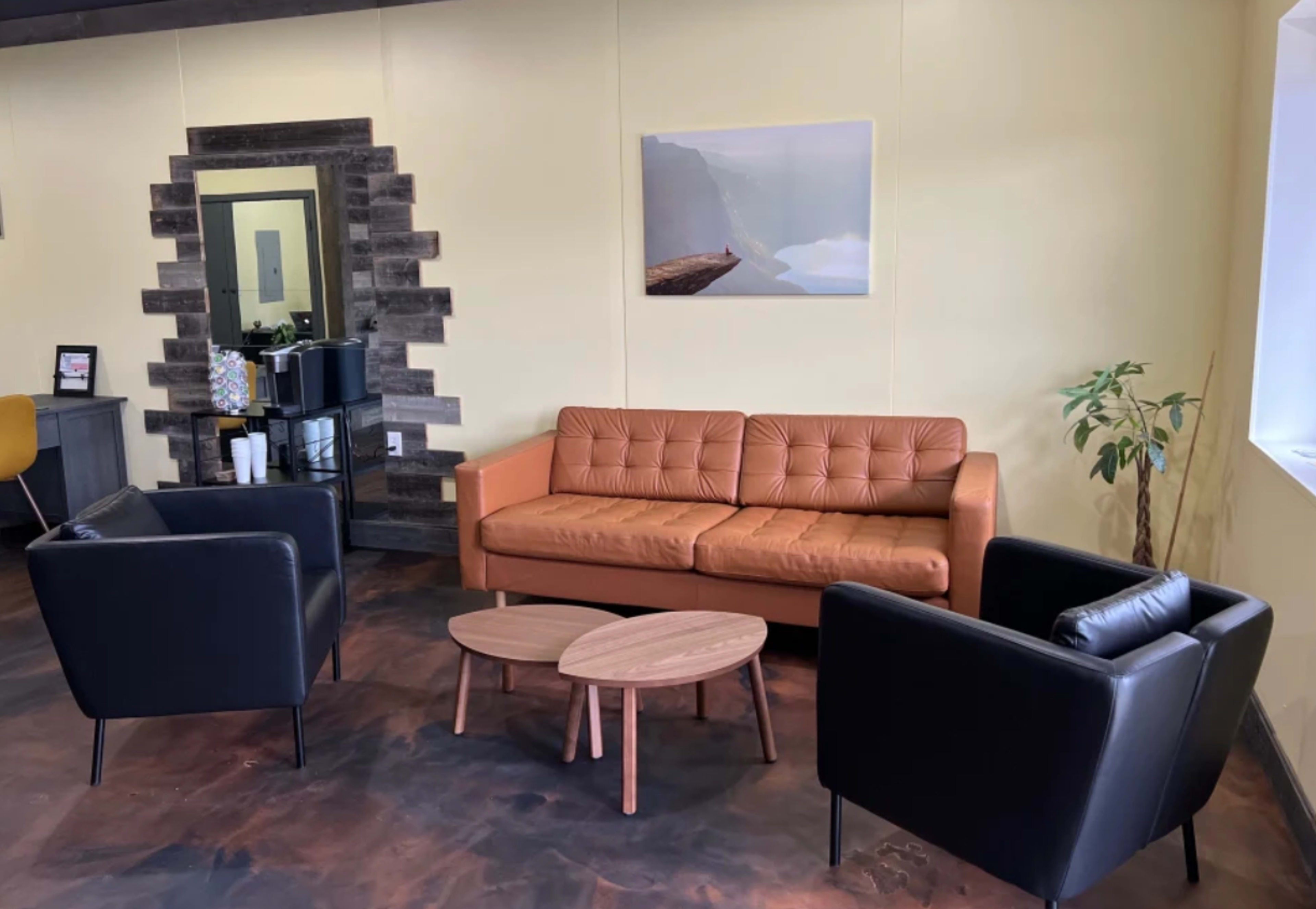 A modern seating area with a brown leather sofa, two black armchairs, and two wooden coffee tables against a yellow wall.