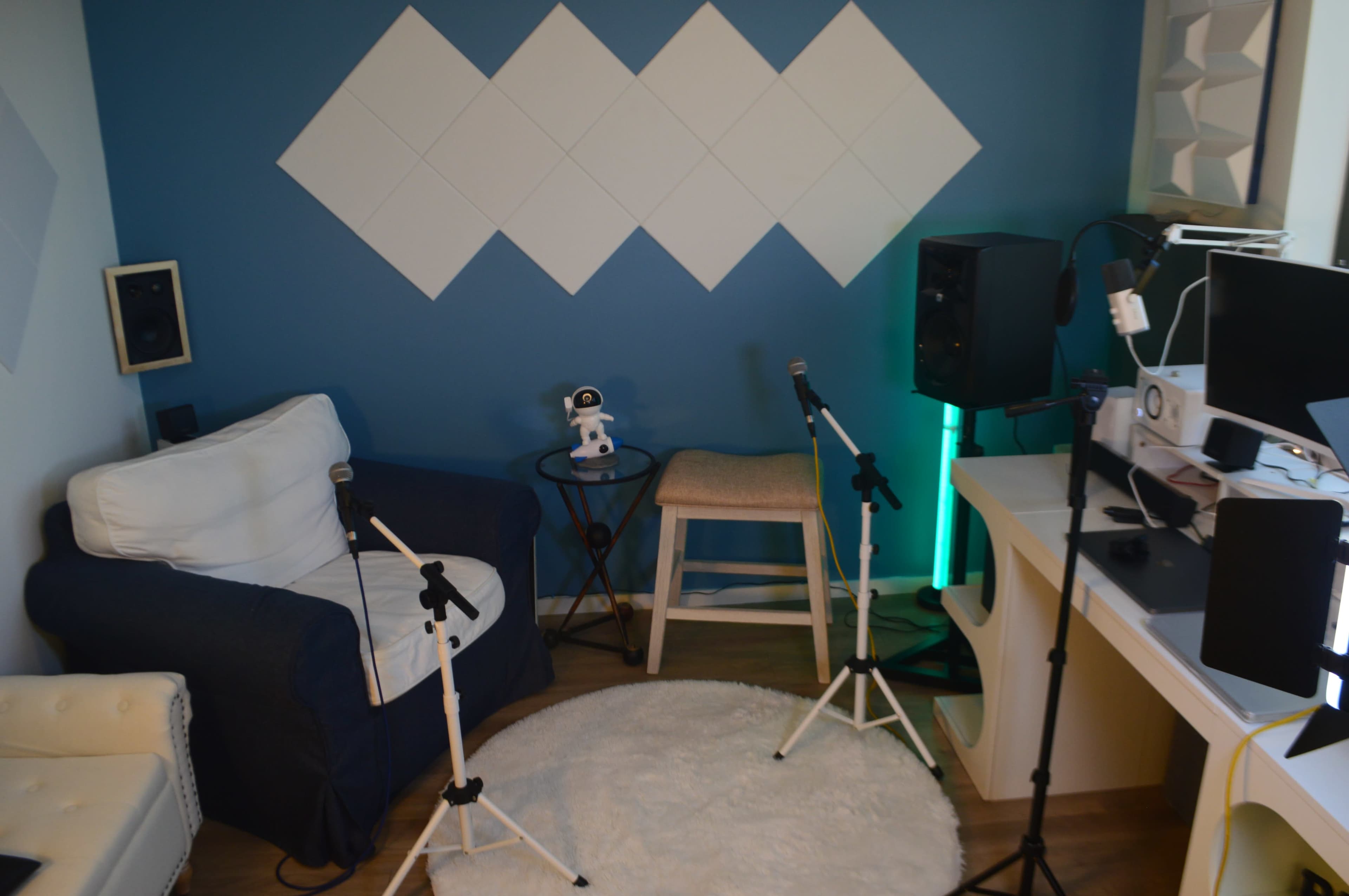 The image shows a small studio space featuring a blue wall with diamond-shaped decorations, two microphones on stands, a stool, a round table, and a desk with electronic equipment.