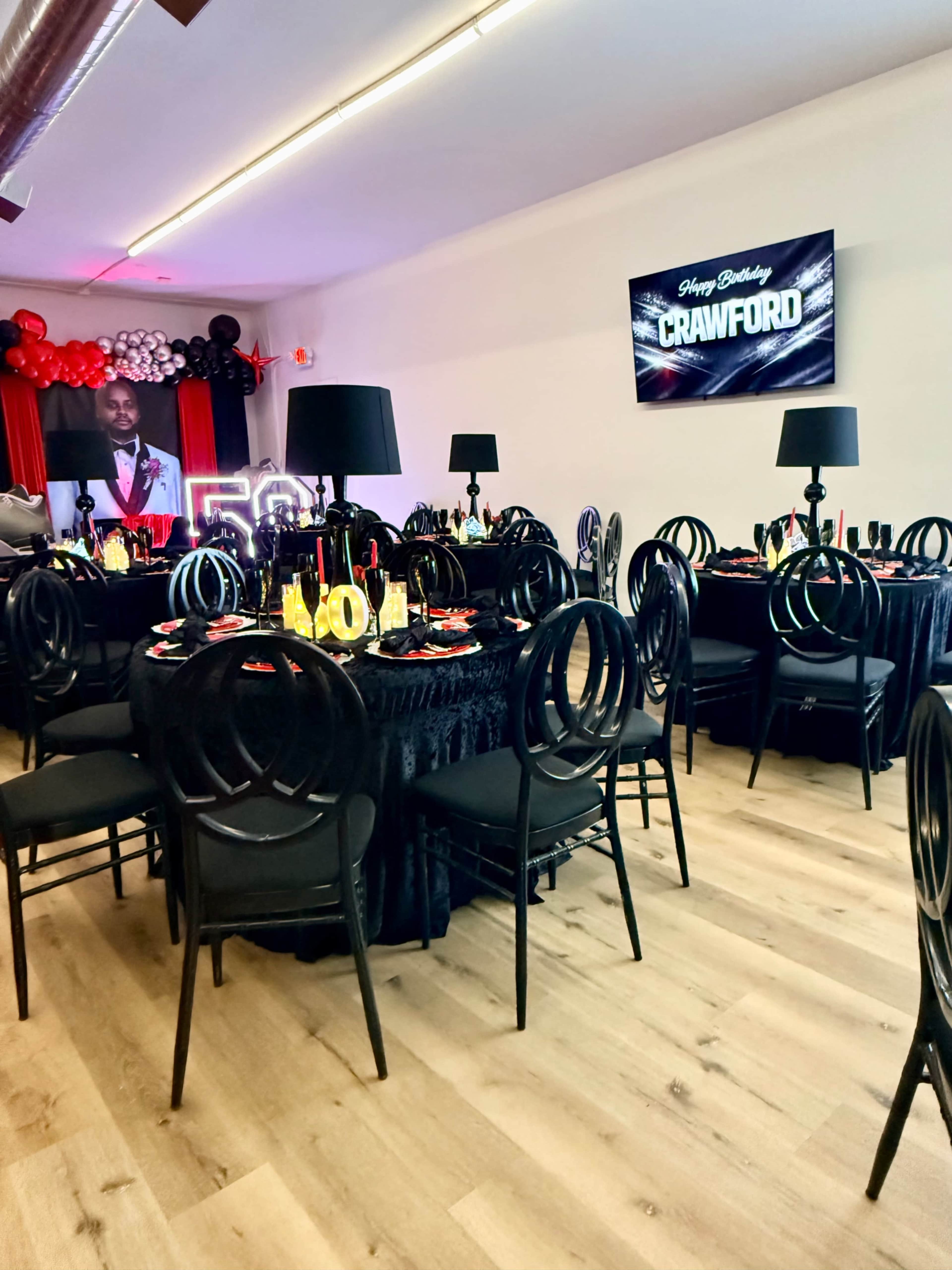 The image shows a decorated party venue with black tables and chairs, red and black balloon arrangements, and a digital screen displaying a birthday message.