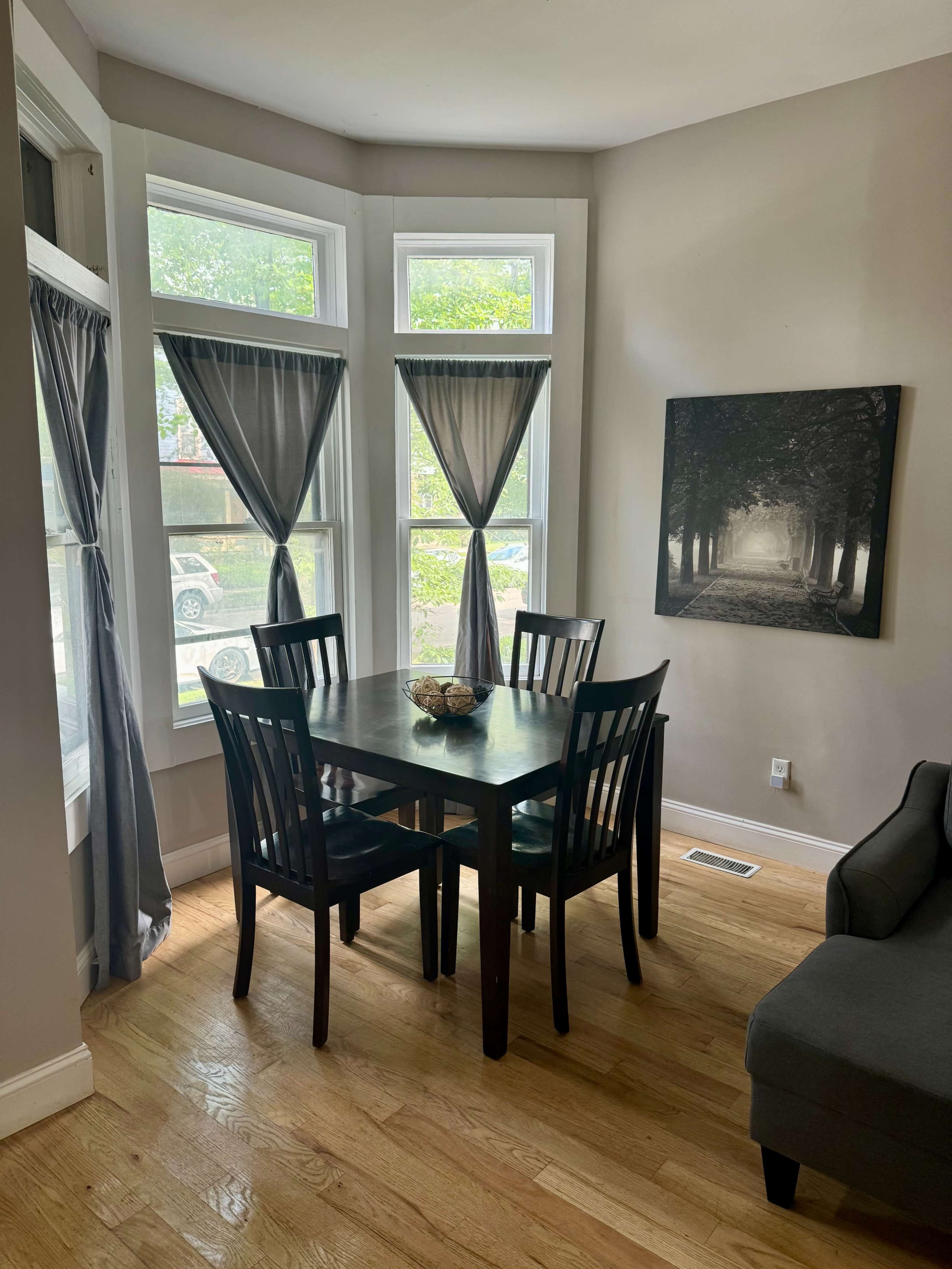 A small dining area features a dark wooden table surrounded by four chairs, situated near large windows with grey curtains.