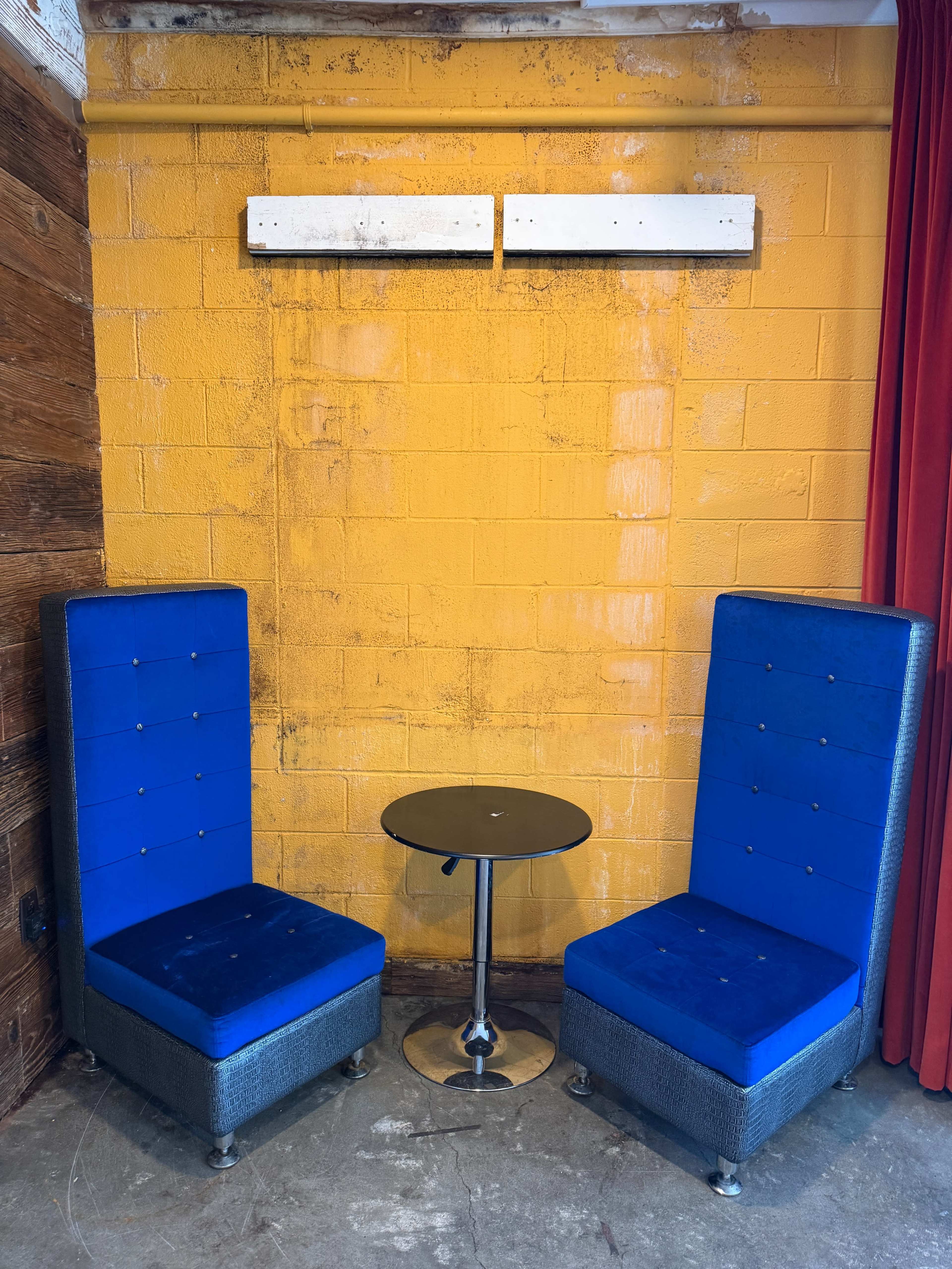 The image shows a small seating area with two blue chairs and a round black table against a yellow wall.