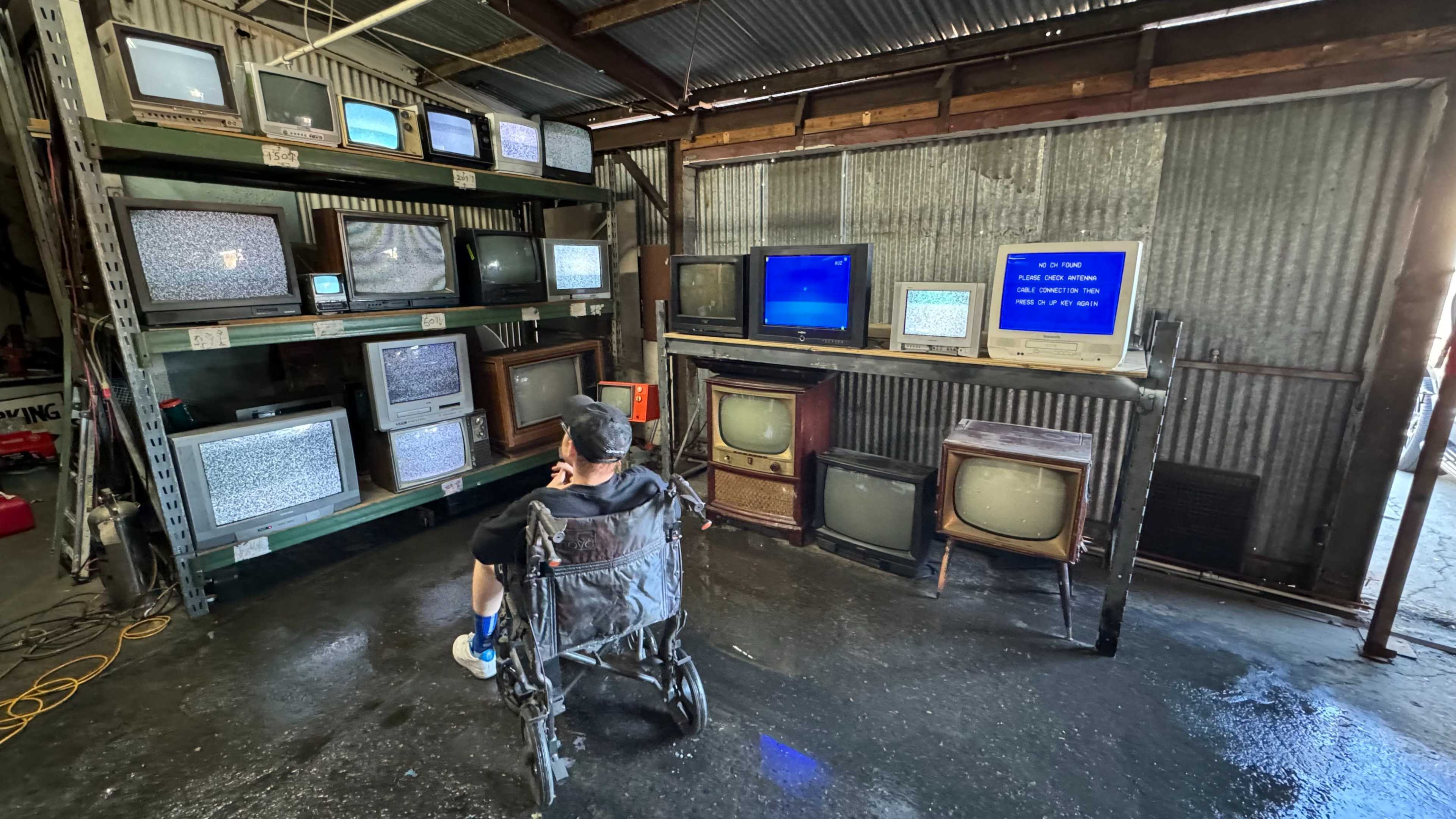 A person in a wheelchair sits in front of multiple old televisions displaying static in a metal-walled room.