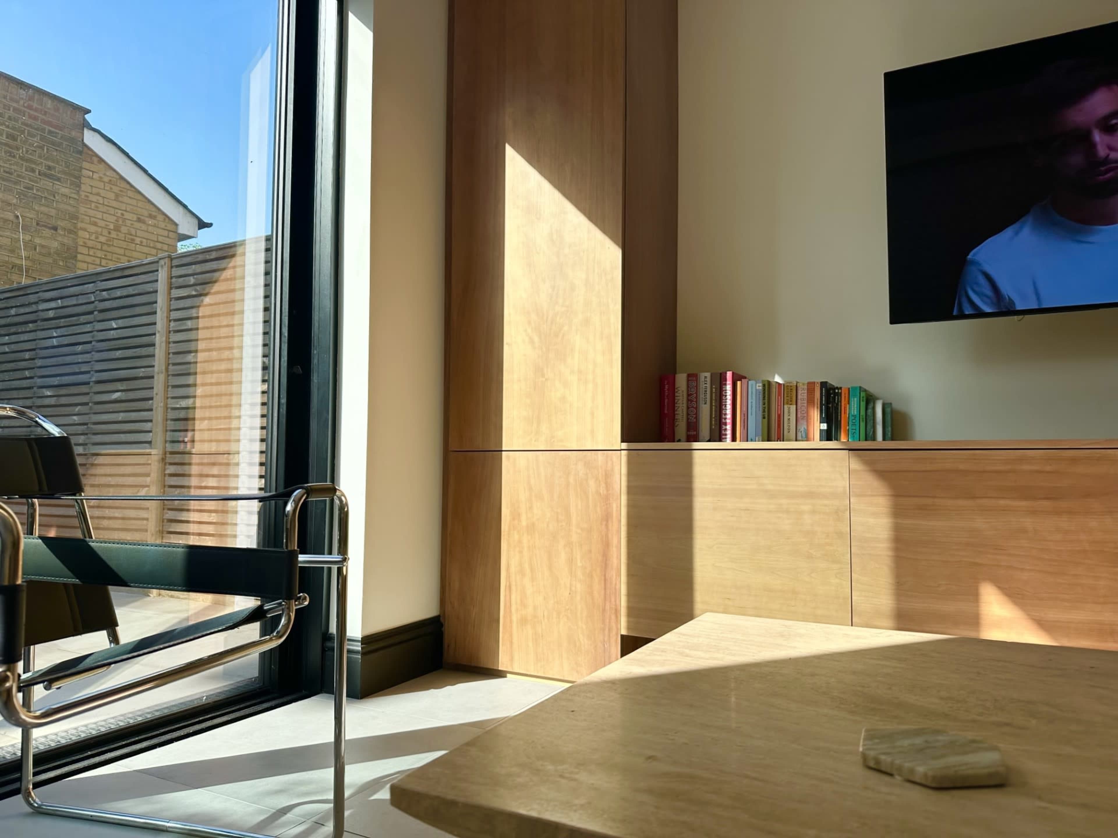 A sunny living room features a wooden cabinet, a television mounted on the wall, and a glass door leading to an outdoor area.