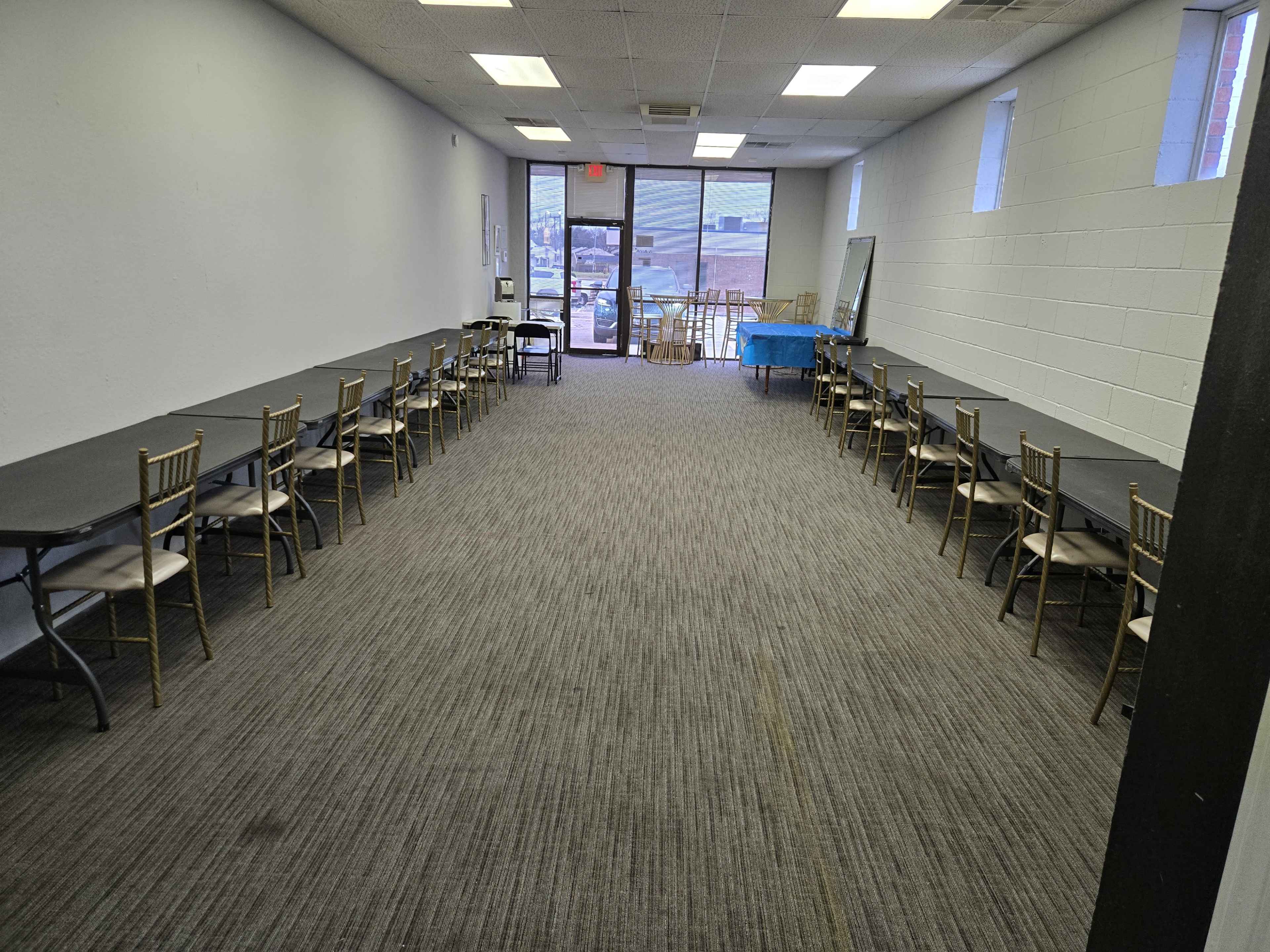 The image shows a long, empty room with two rows of tables and chairs, leading to a doorway that opens to an outdoor area.