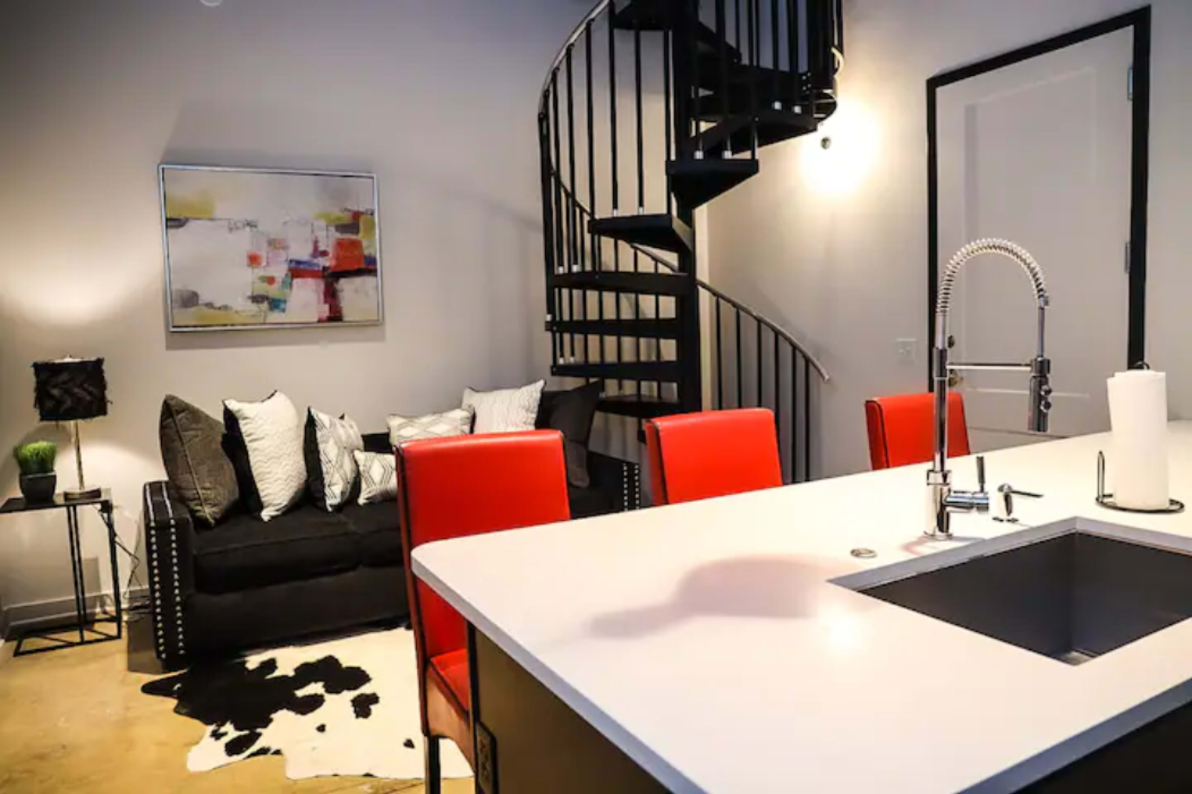 A modern living space featuring a spiral staircase, a black sofa with decorative pillows, a white kitchen island with red chairs, and abstract wall art.