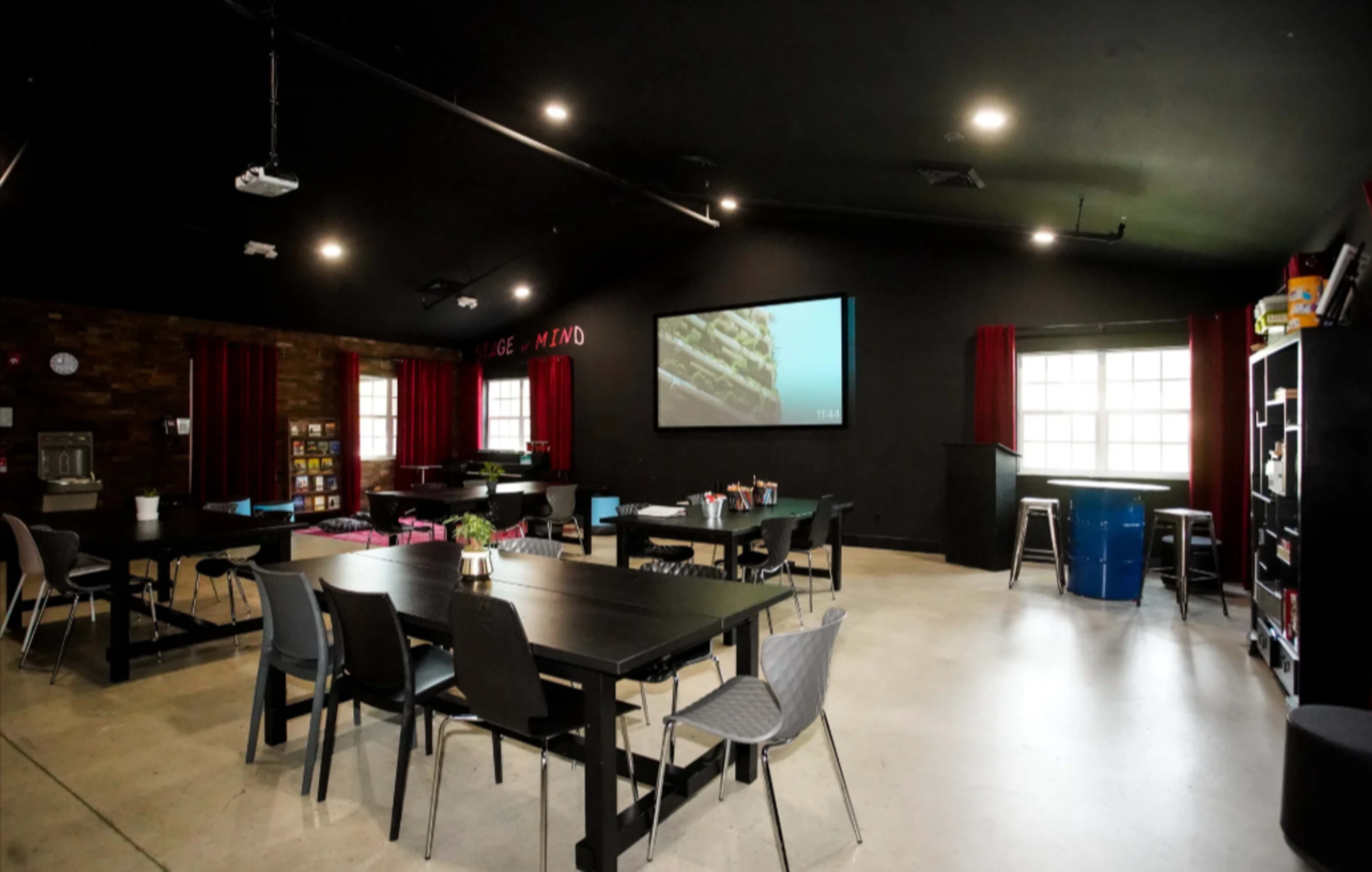 The image shows a spacious room with a black ceiling and walls, featuring several tables, chairs, a projection screen, and a large window with red curtains.