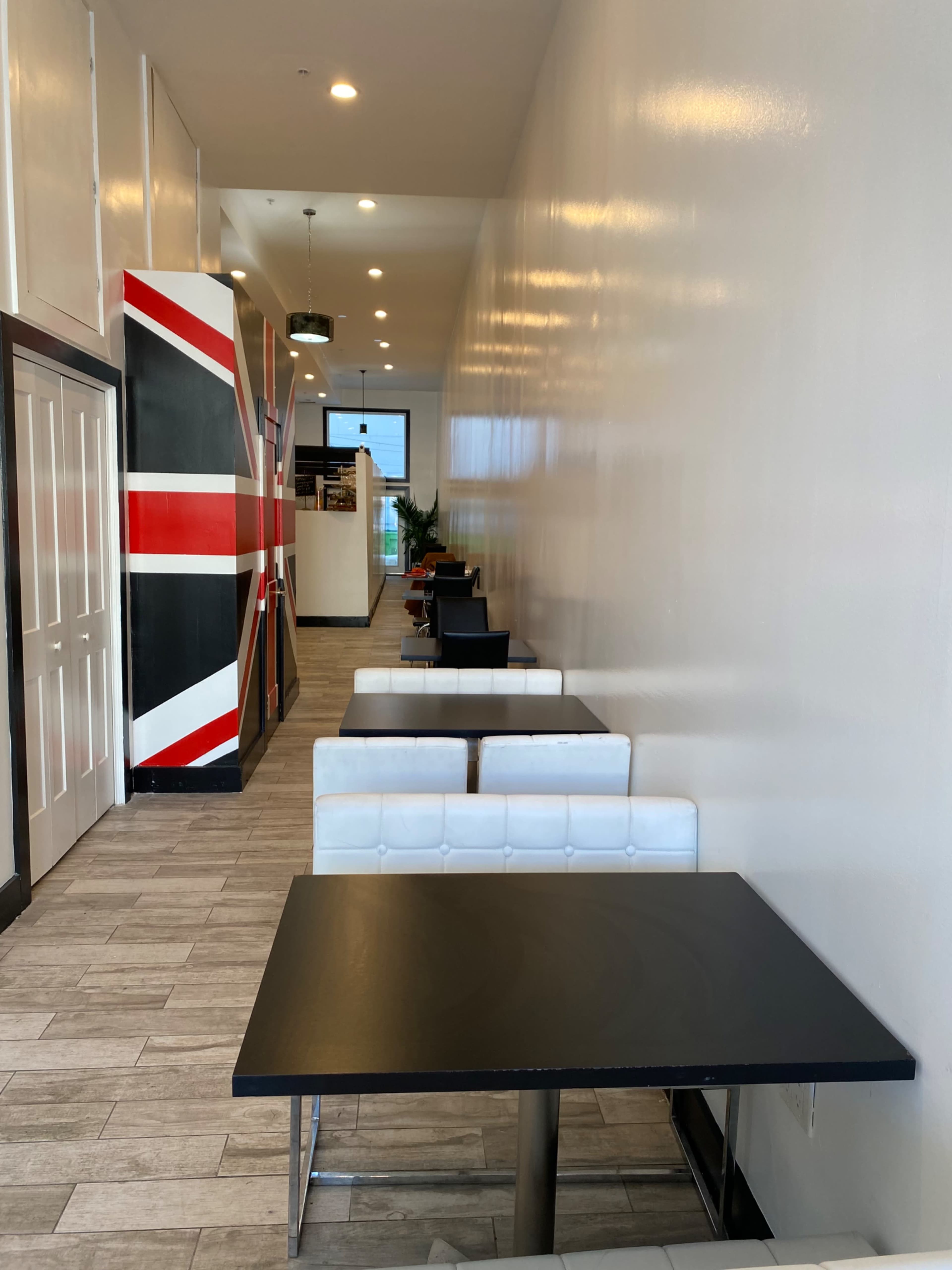 A narrow, well-lit hallway in a café, featuring black tables with white chairs along the sides and a large painting of the Union Jack on the wall.