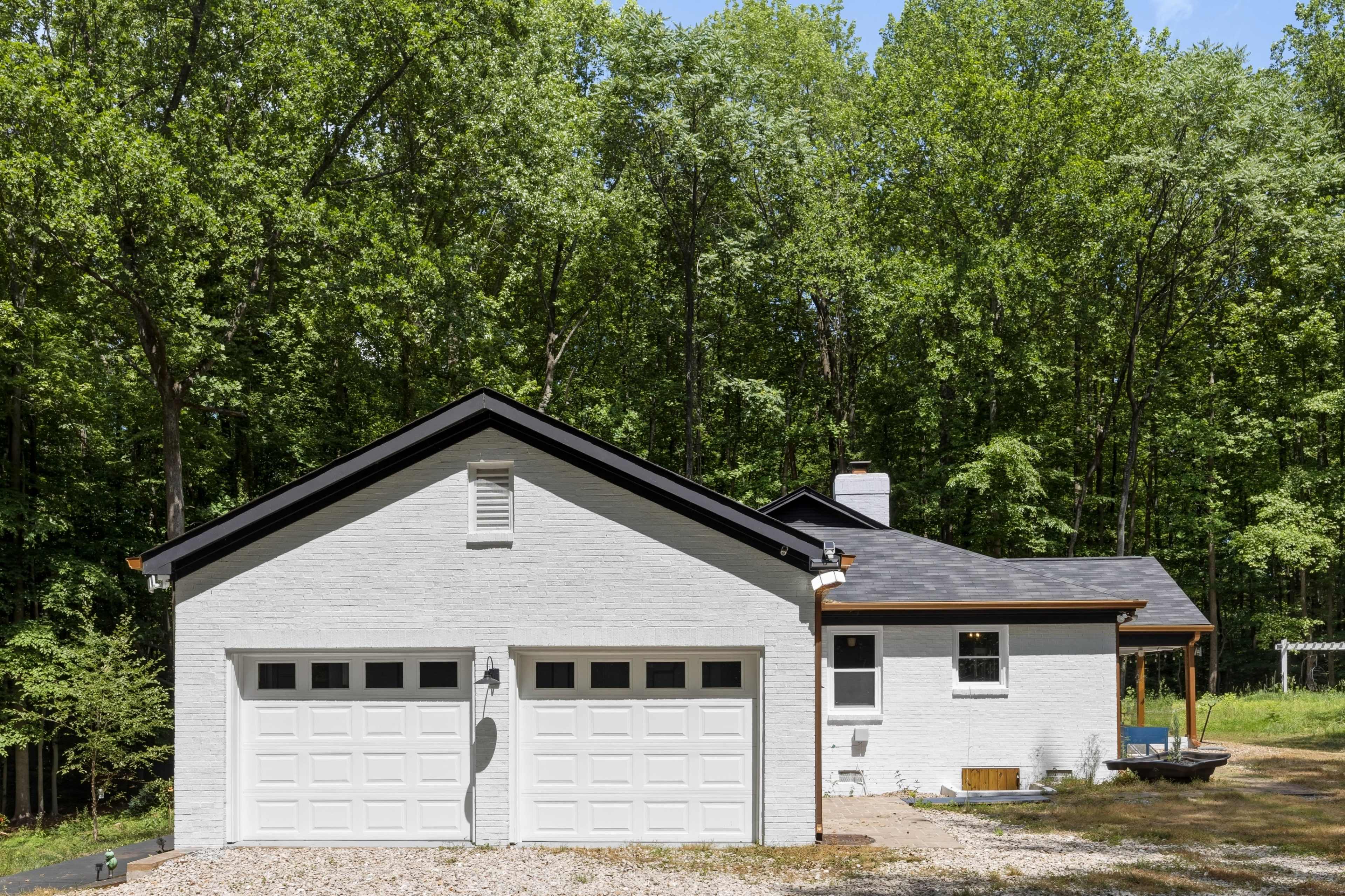 A white brick house with a two-car garage is surrounded by lush green trees.