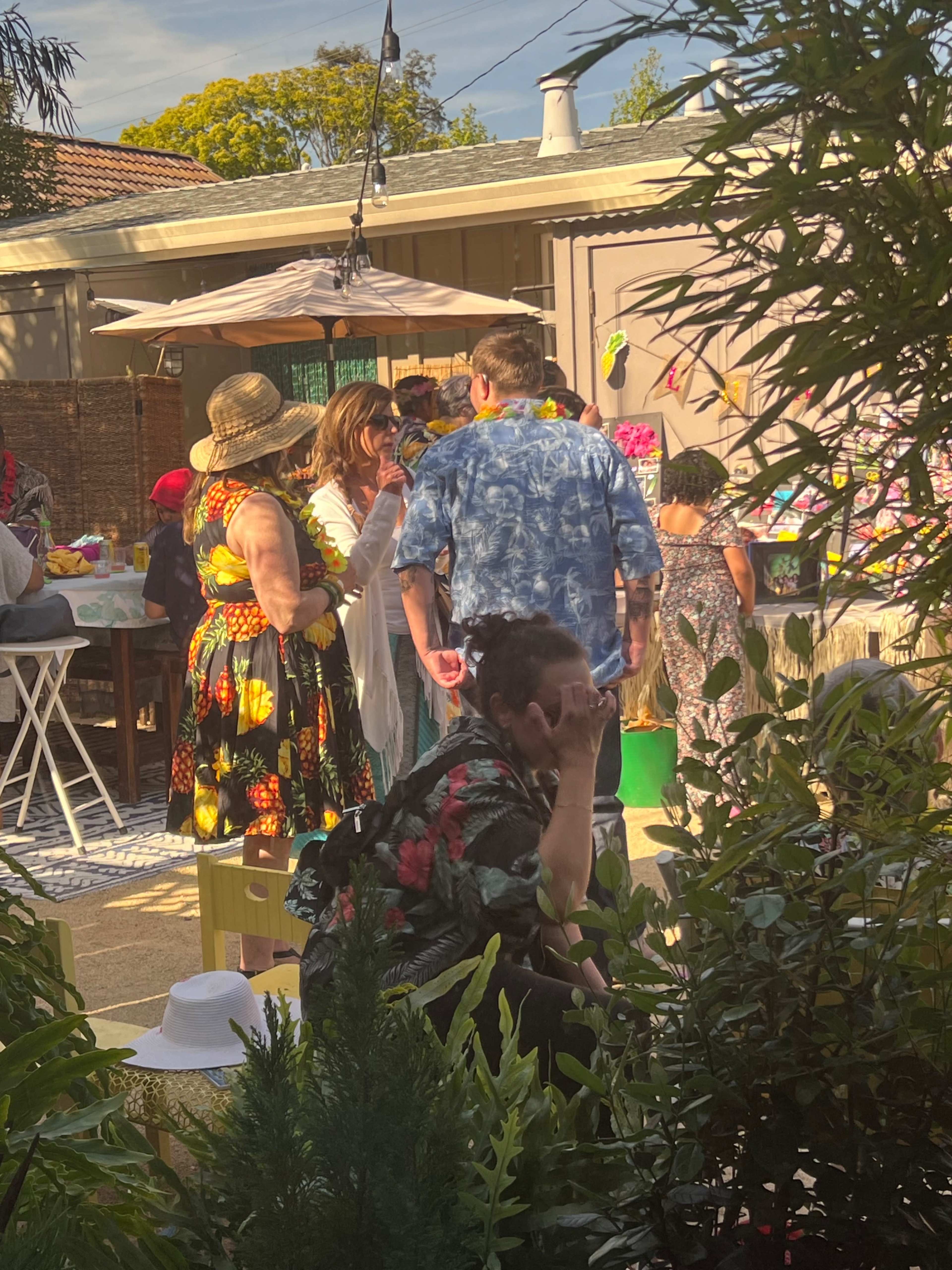 A group of people gathers in a backyard decorated for a celebration, with tables set up and greenery surrounding the scene.