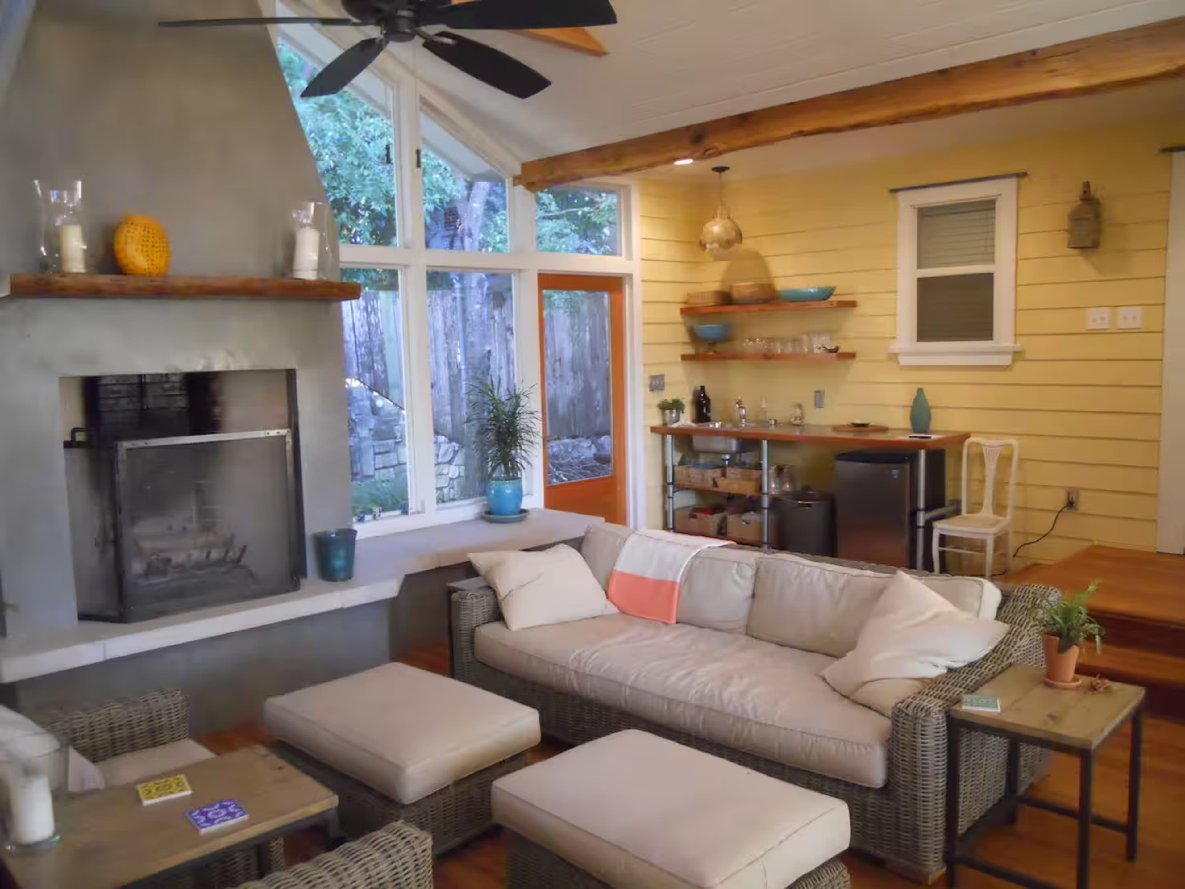 The image shows a modern living room with a fireplace, a sectional sofa, and a small dining area with a bar cart, all illuminated by natural light from large windows.