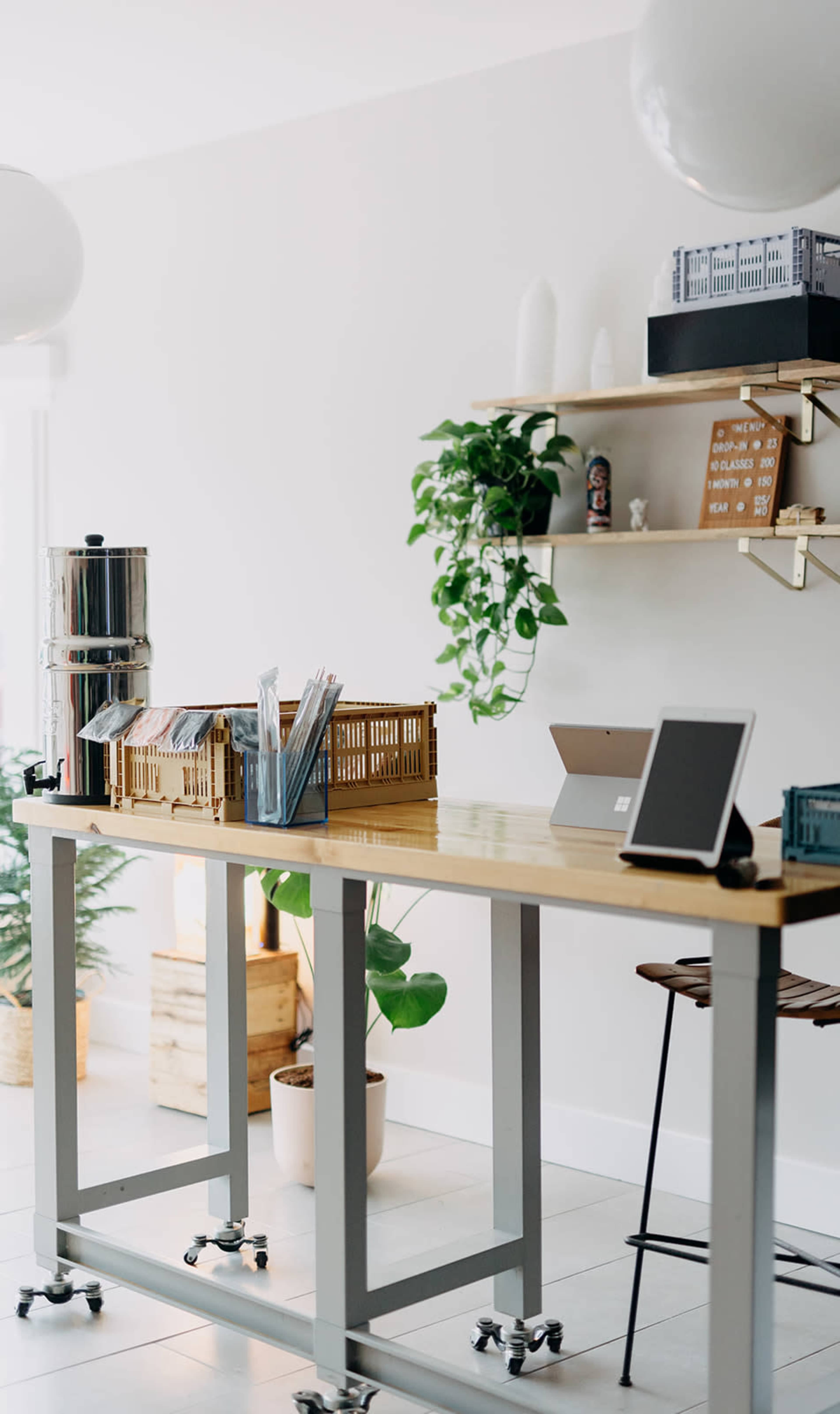 A modern workspace features a long wooden table with a beverage dispenser and organizational baskets, against a backdrop of greenery and minimalist decor.