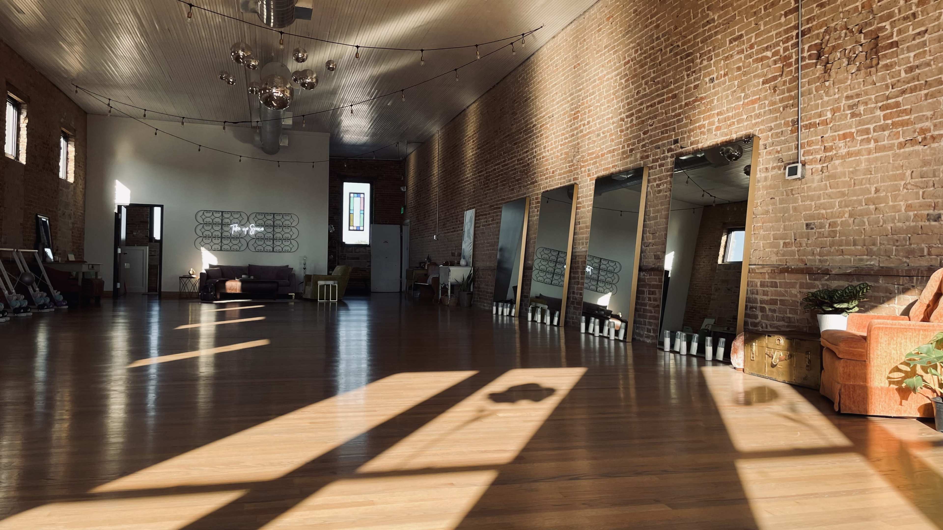 The image shows a spacious room with exposed brick walls, large mirrors, and sunlight casting long shadows on the wooden floor.
