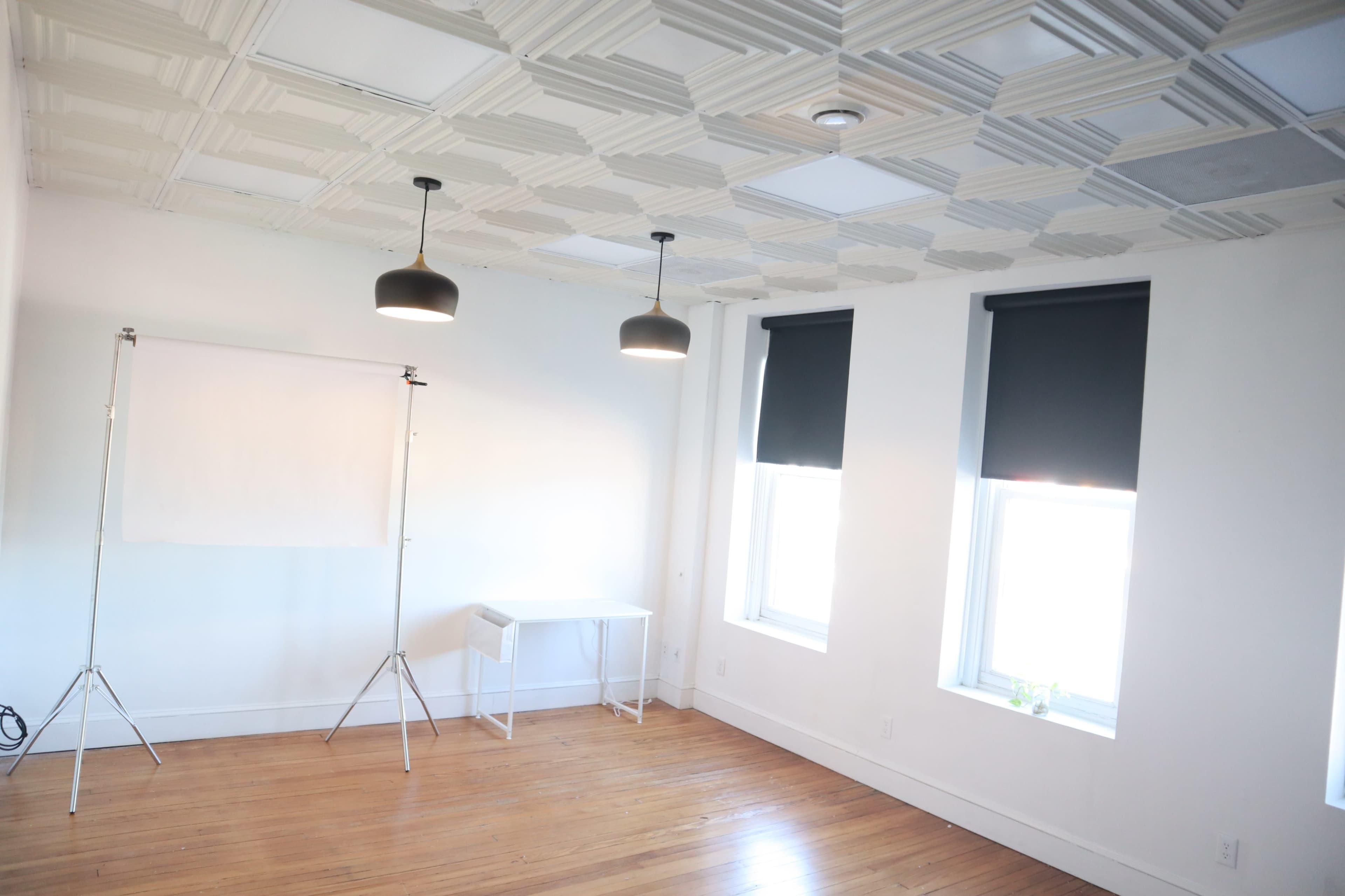 A bright, minimalist room with a patterned ceiling, two windows fitted with black roller shades, and a small white table beside a backdrop stand.
