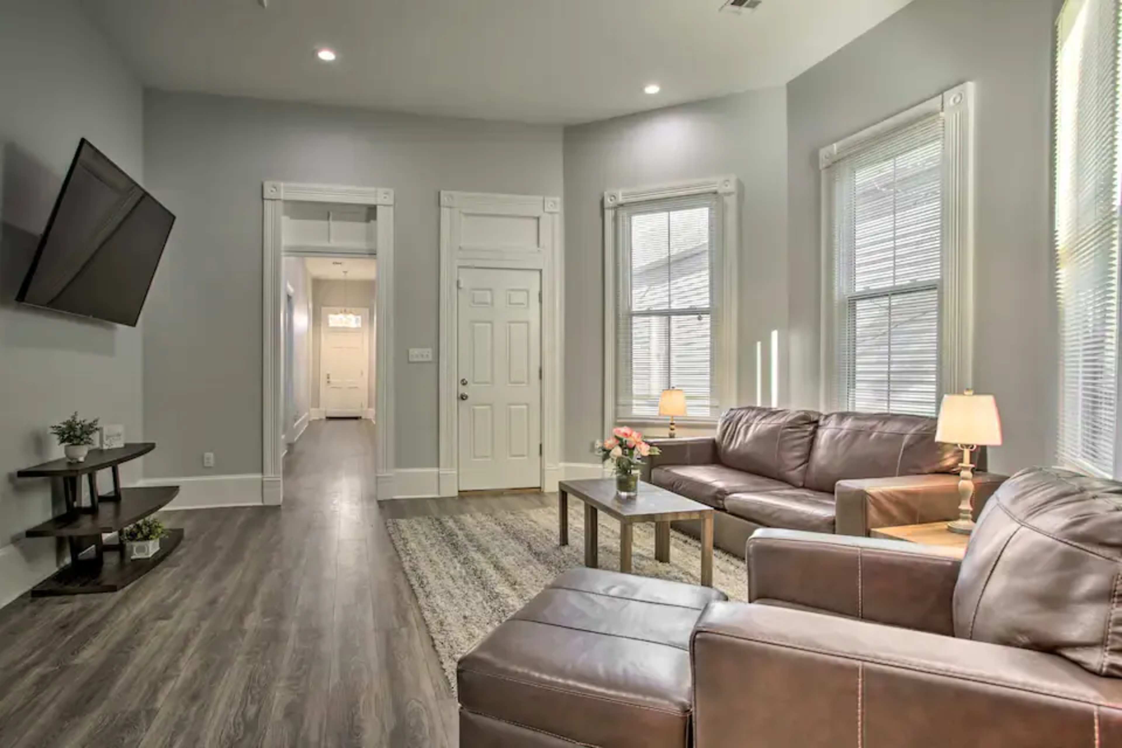A modern living room featuring two brown leather sofas, a coffee table, a wall-mounted TV, and bright windows, with a hallway visible in the background.