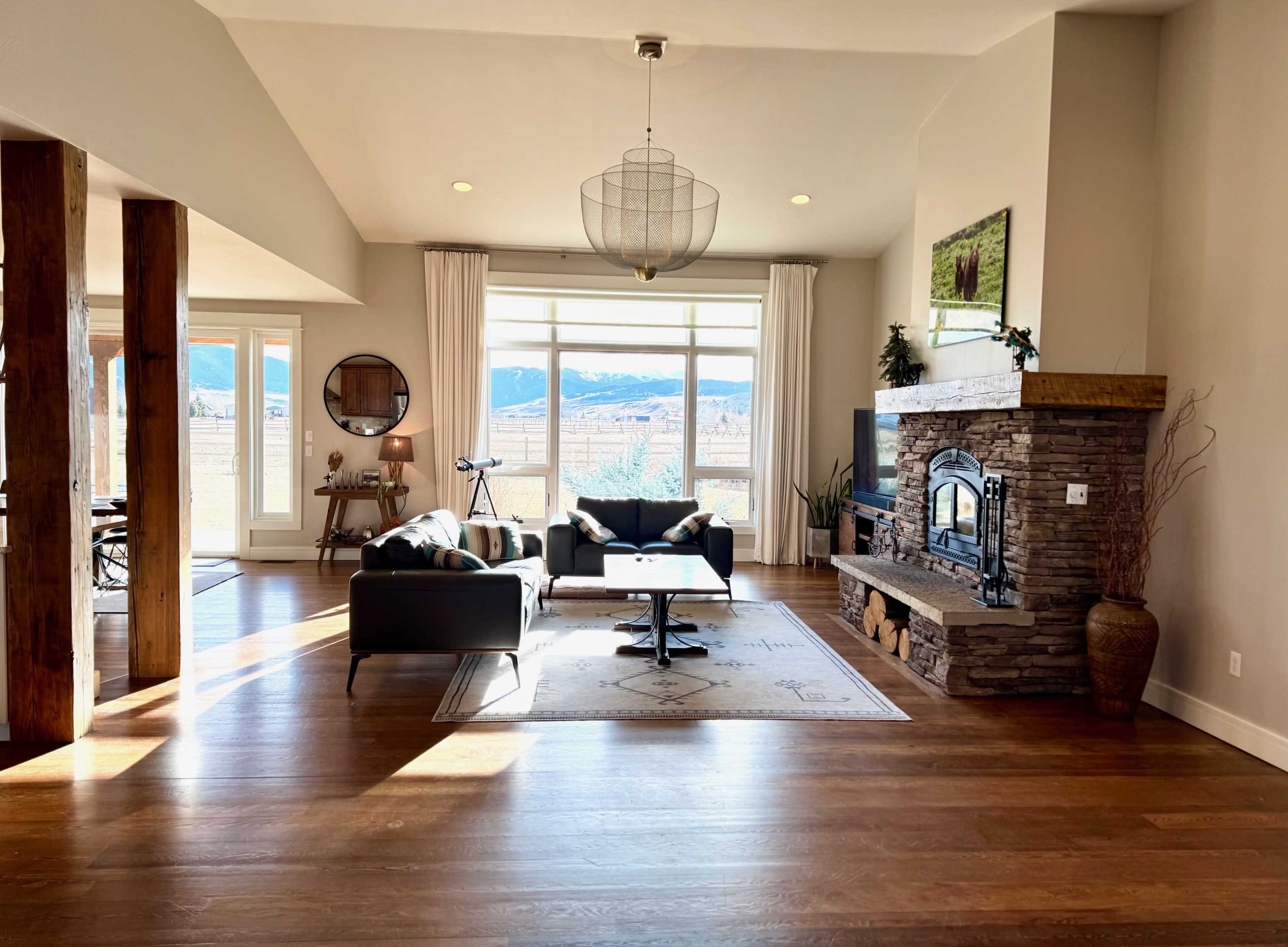The interior of a spacious living room features a stone fireplace, large windows with mountain views, and modern furniture.