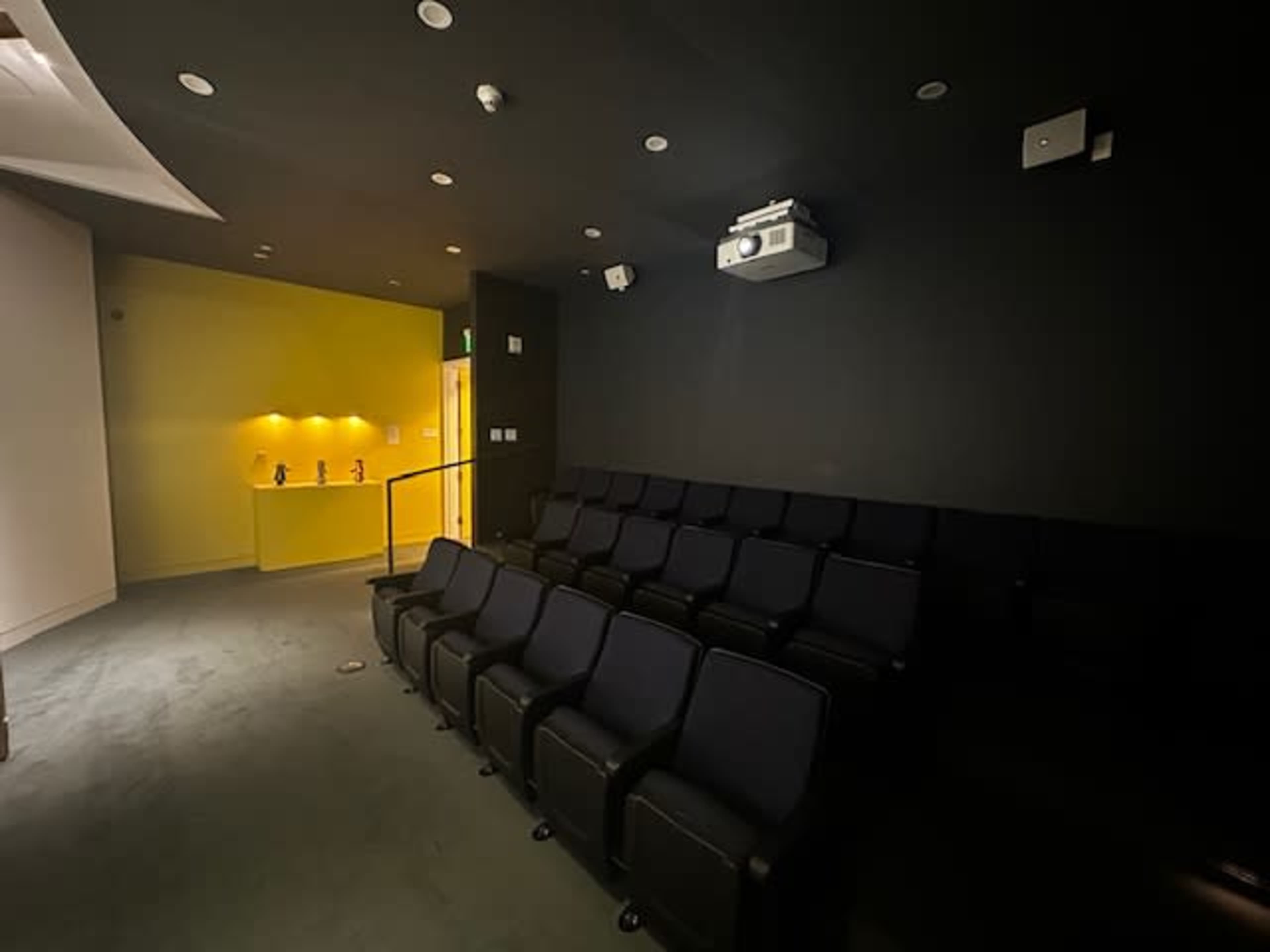 A small, dark theater room with several rows of black seats facing a projector screen and a yellow-lit wall.
