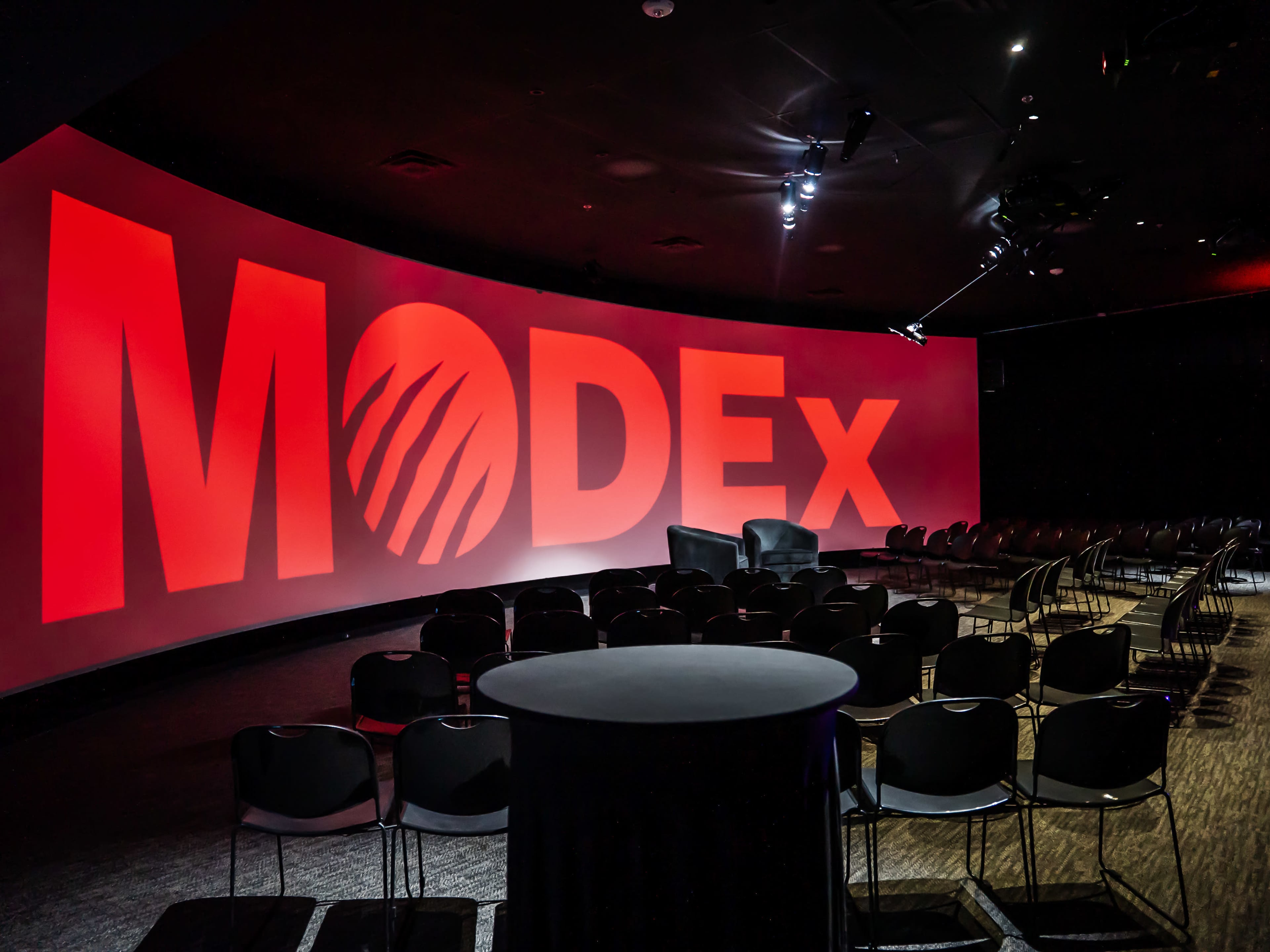 A large auditorium features a red backdrop with the word "MODEX" prominently displayed, arranged with chairs facing a central table.