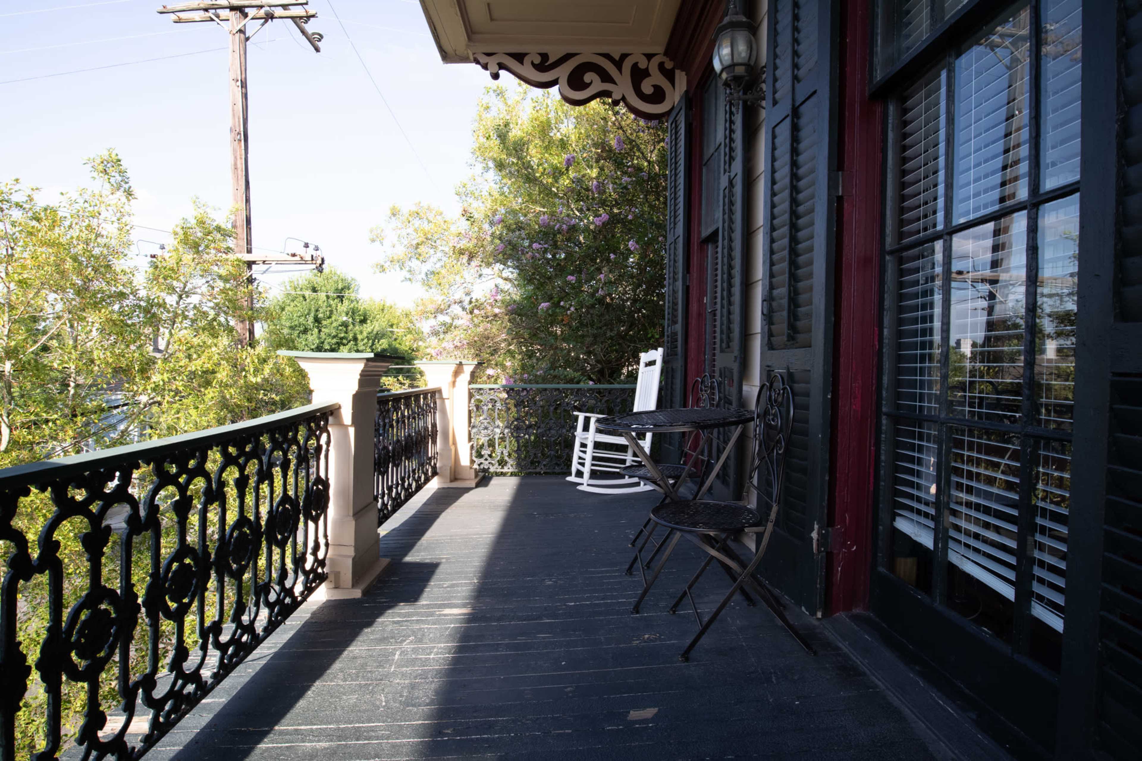 The image shows a balcony with a rocking chair and a small table, surrounded by a wrought iron railing and greenery in the background.