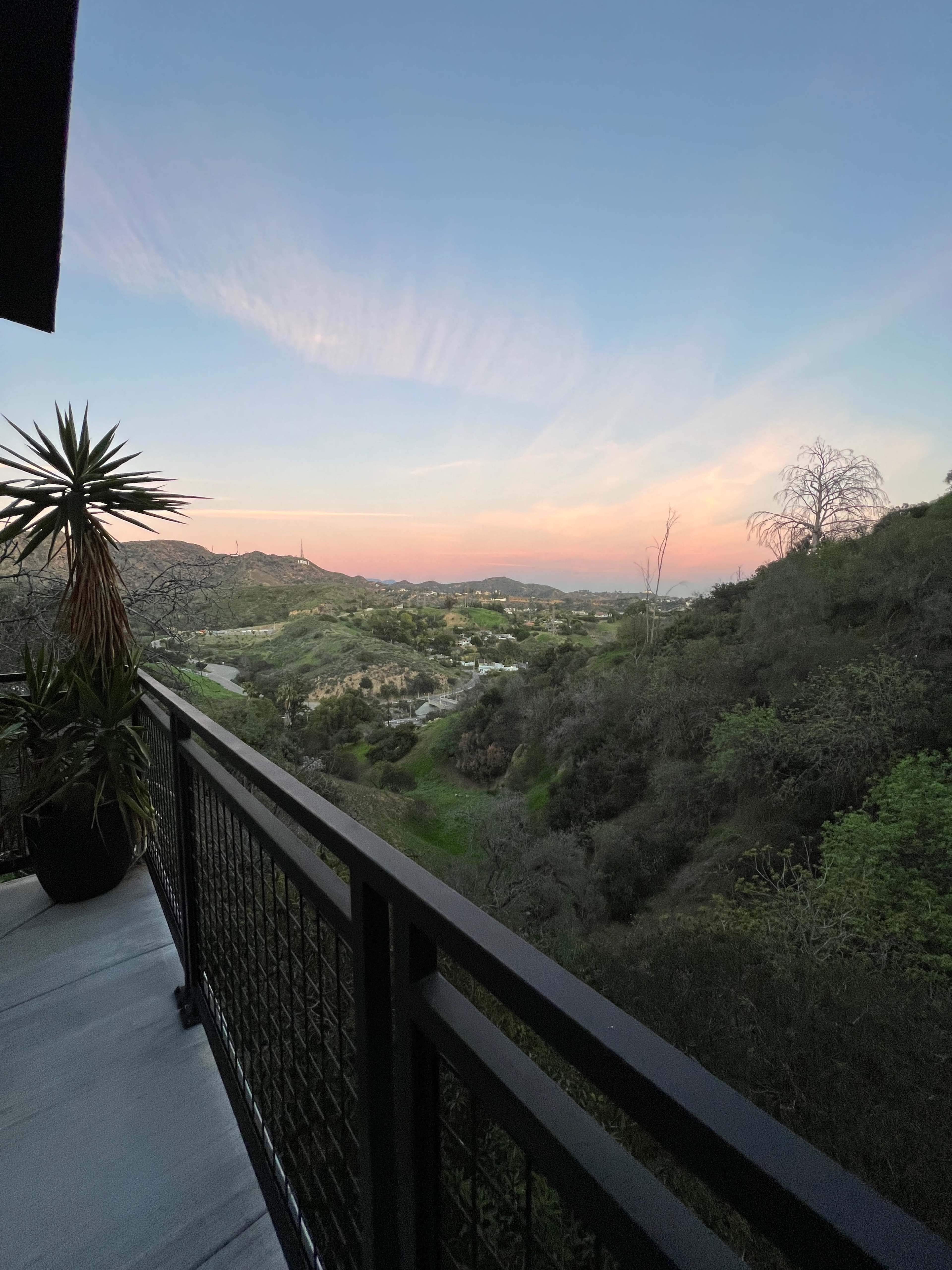 A balcony overlooks a hilly landscape during sunset, with a mix of greenery and distant hills under a colorful sky.