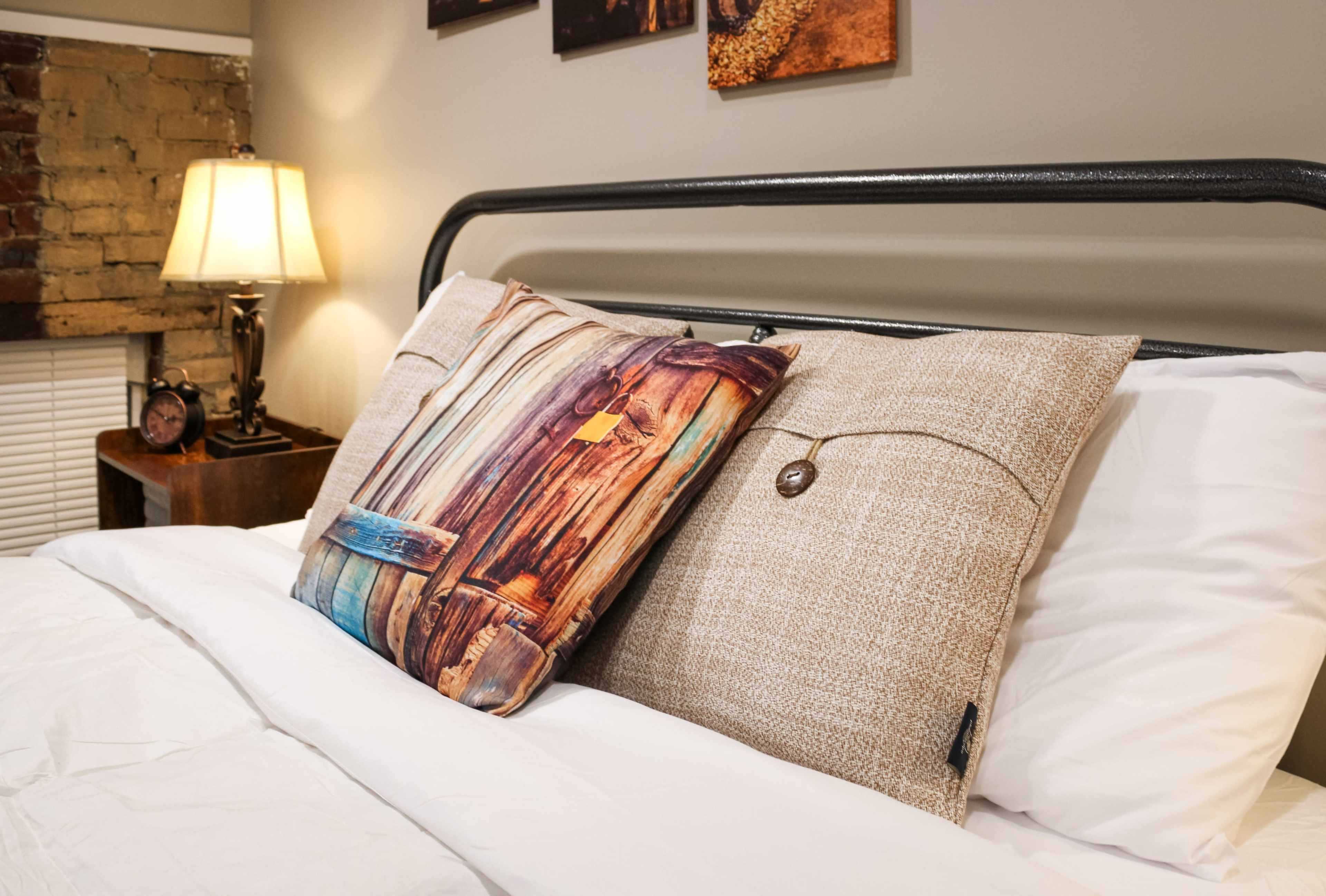 A neatly made bed features two decorative pillows against a backdrop of a bedside lamp and framed photographs on the wall.