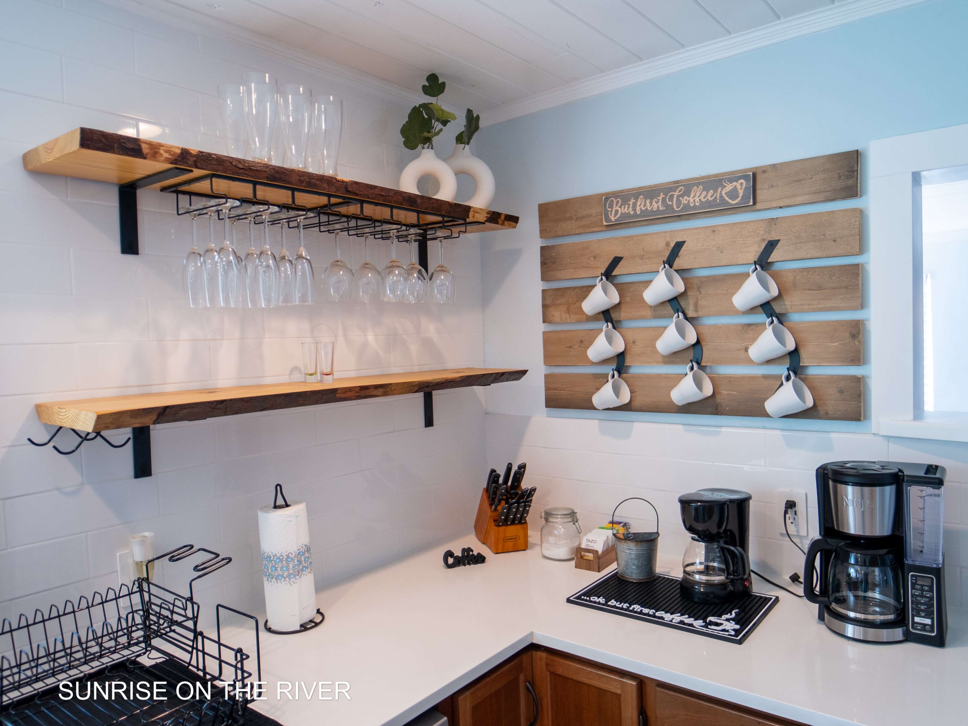 A kitchen corner with two wooden shelves displaying glassware and a wall-mounted rack holding white mugs, alongside a coffee maker and utensils on the counter.