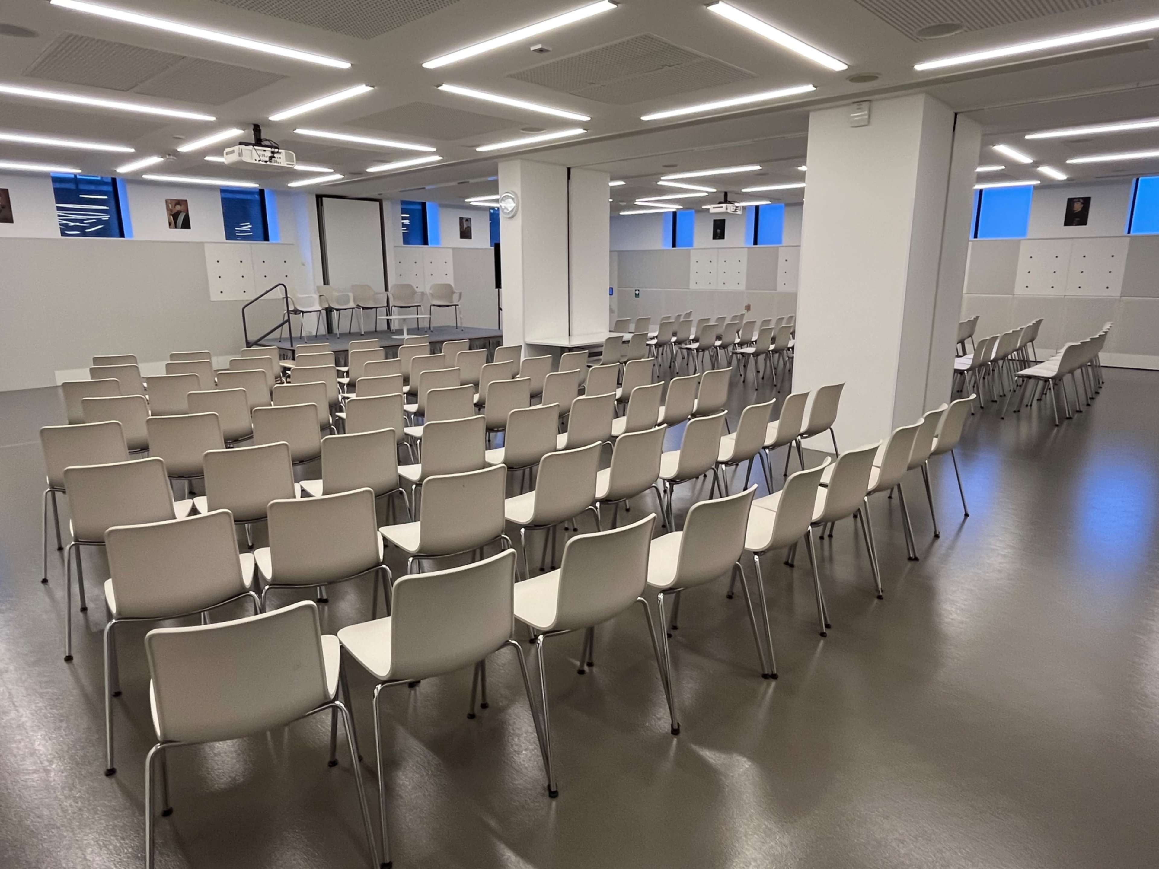 The image shows a modern conference room set up with rows of beige chairs arranged for an audience, and a presentation space visible at the front.