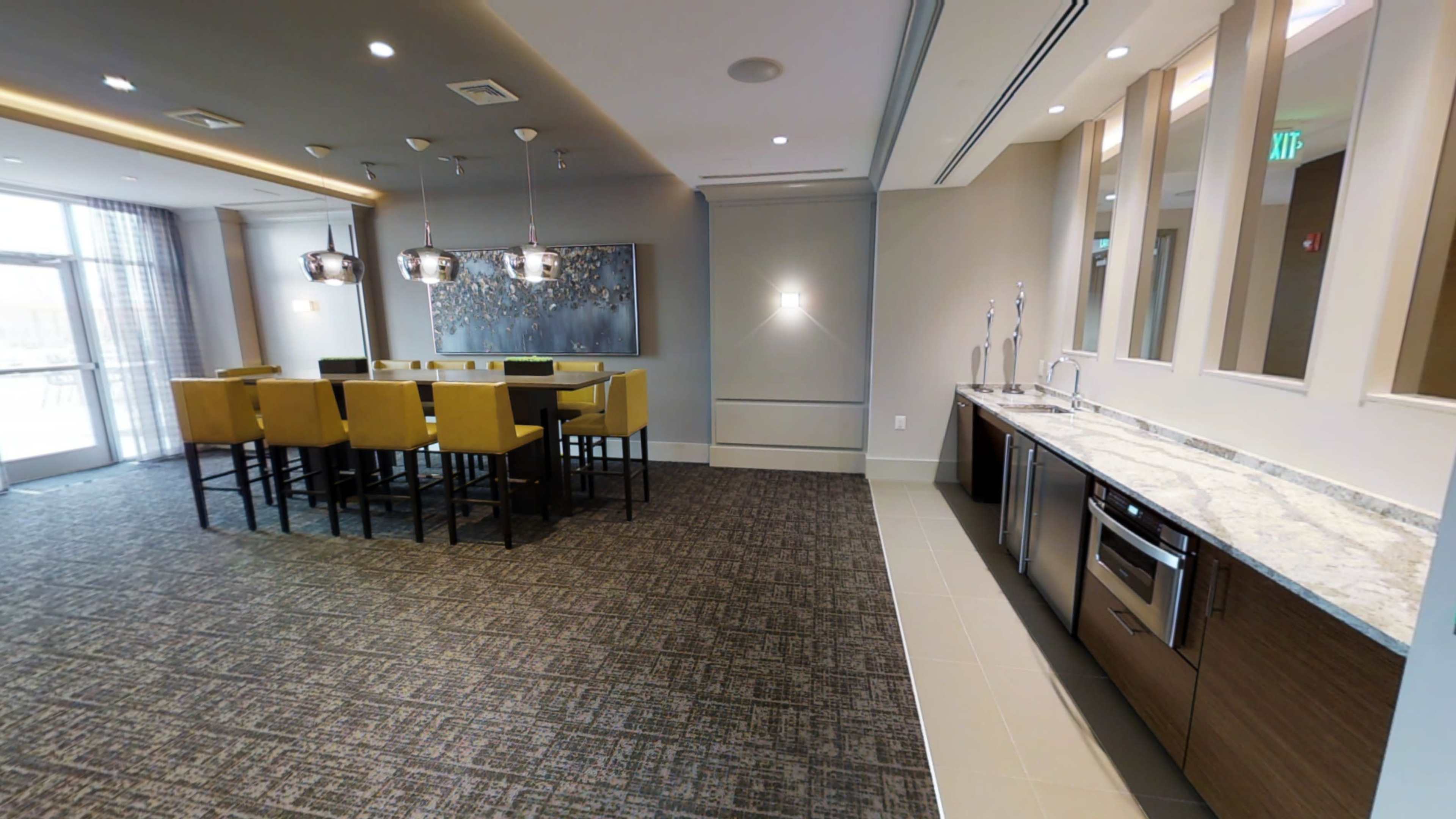 A modern communal kitchen and dining area featuring a long table with yellow chairs, a marble countertop, and wall-mounted appliances against a neutral backdrop.