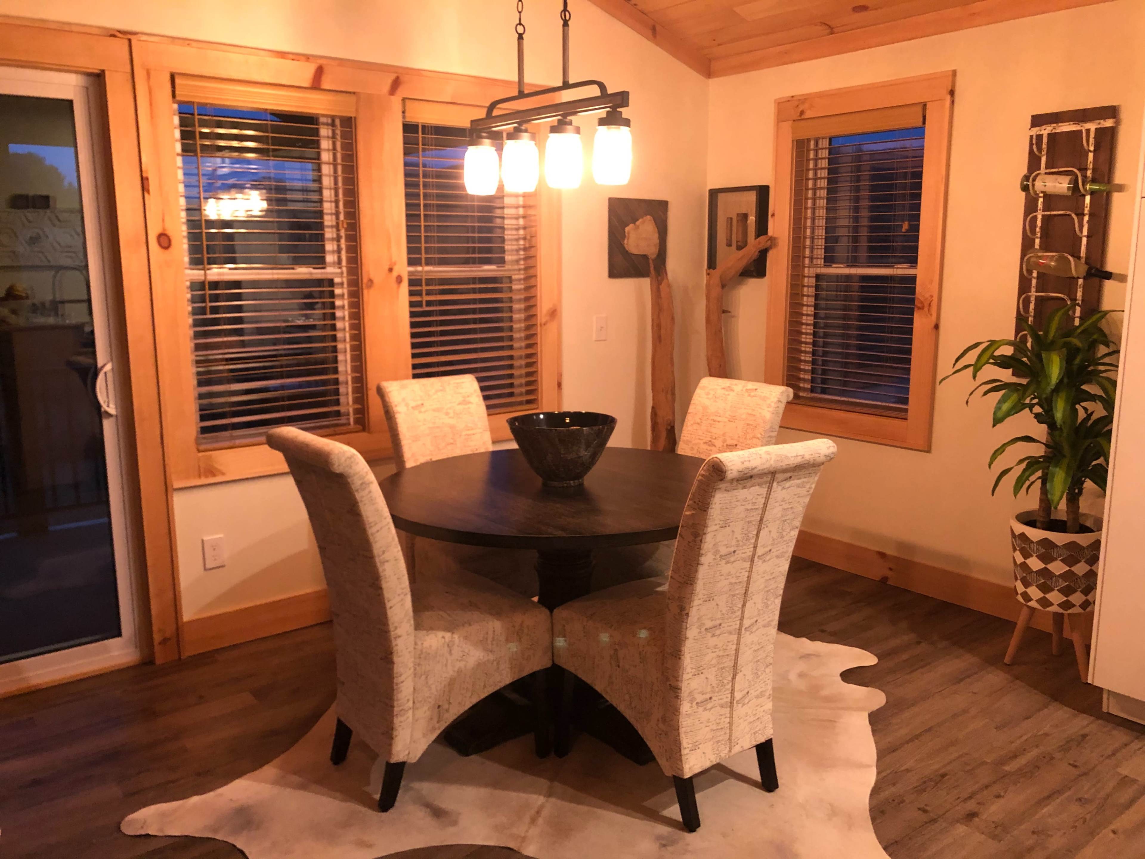 A round dining table with four upholstered chairs is situated in a bright room featuring large windows and wooden accents.