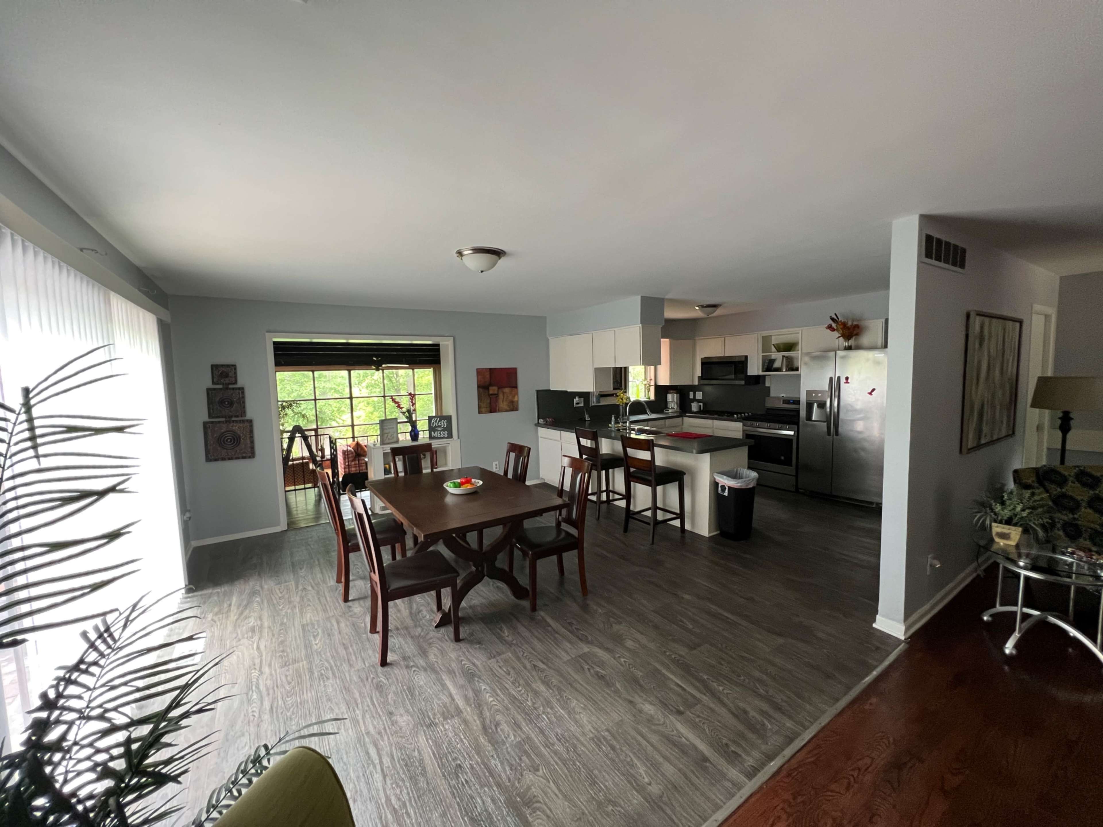 A spacious dining area with a wooden table and chairs next to a kitchen, featuring light gray walls and large windows.