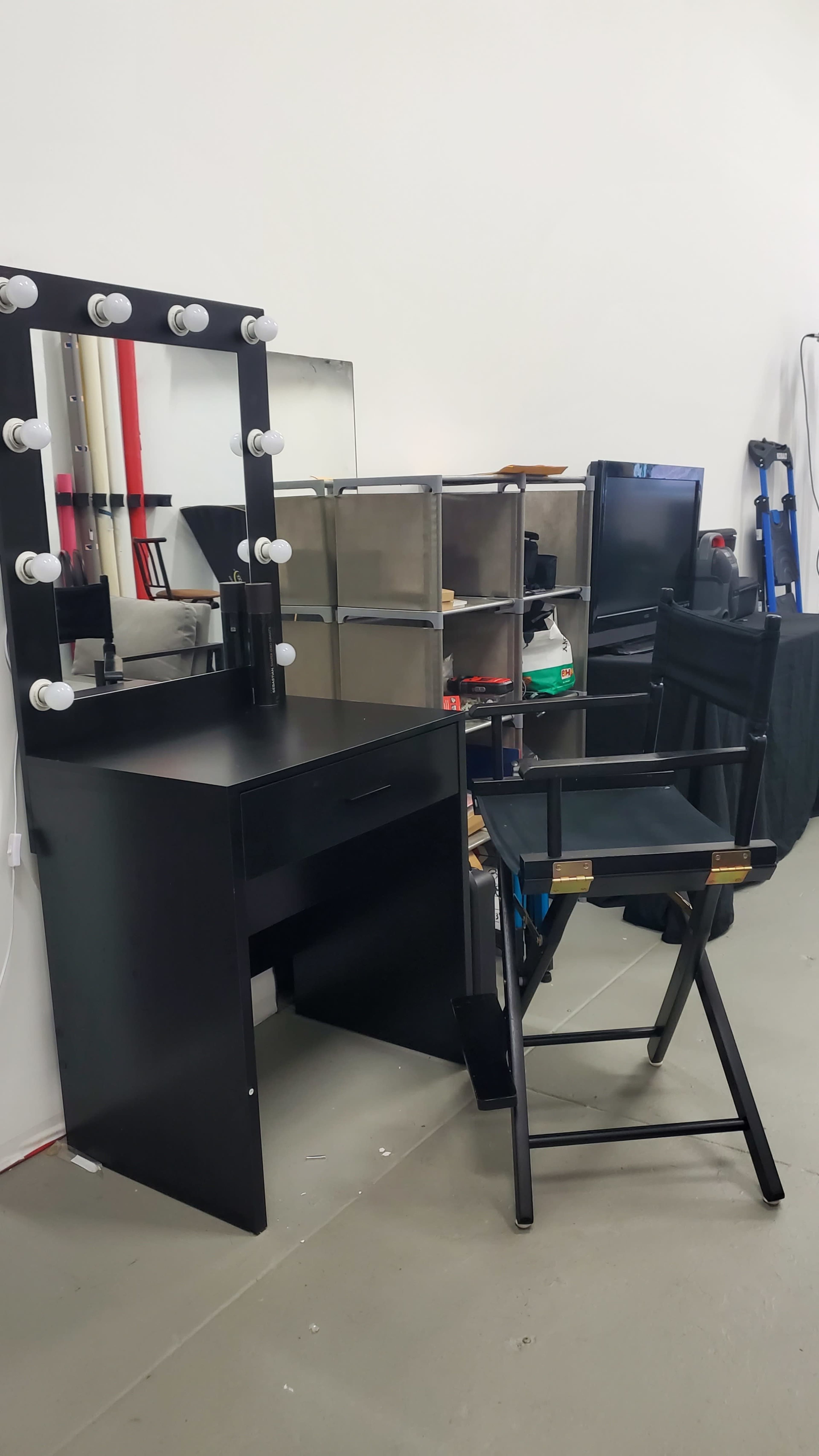 The image shows a black vanity table with lights, a mirror, and a director's chair in a room with storage shelves and various equipment.