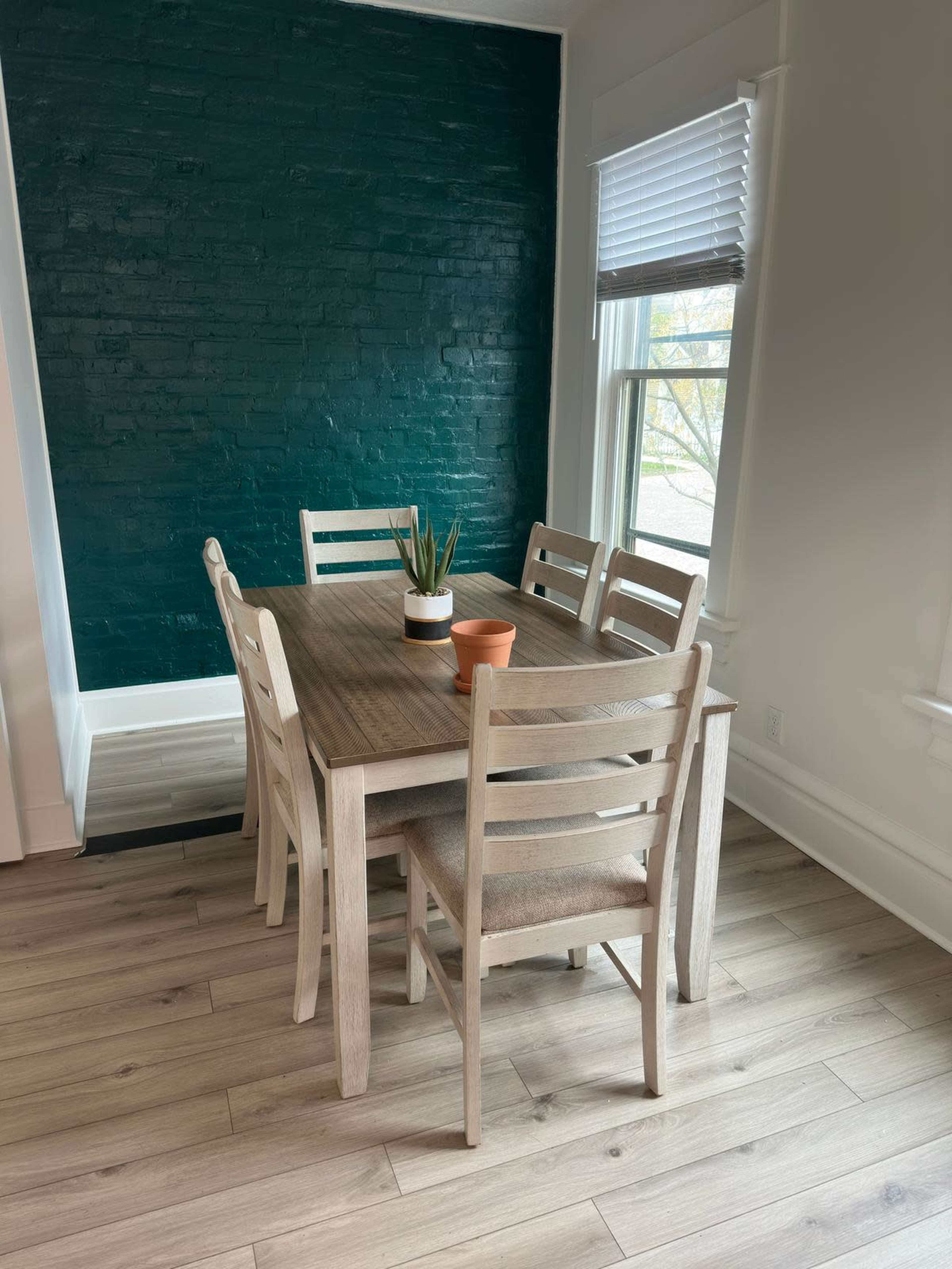 A wooden dining table with four chairs is situated in a brightly lit room, next to a green brick wall and two windows.