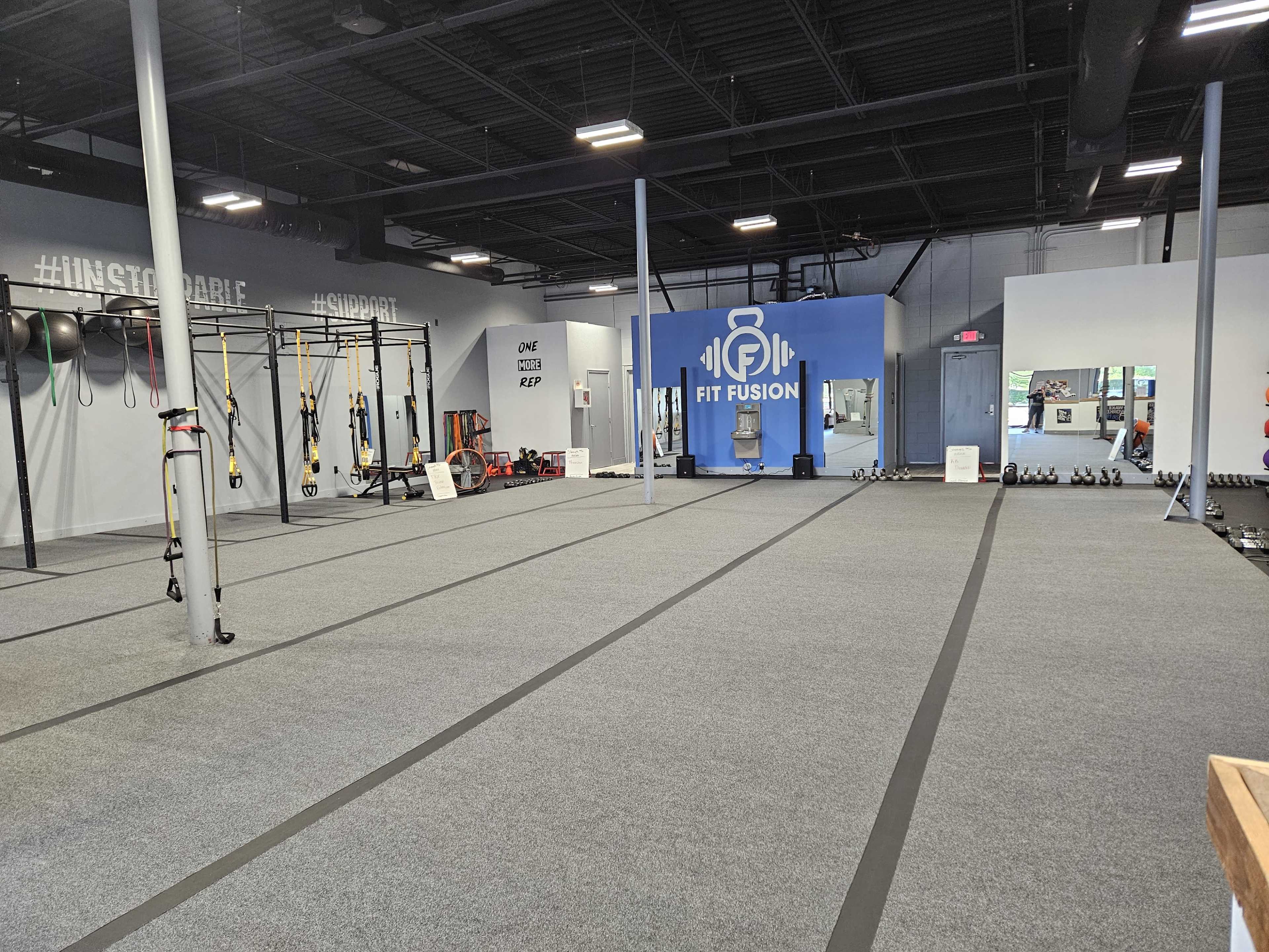 A spacious fitness studio featuring exercise equipment such as kettlebells, TRX suspension trainers, and a large blue wall with the logo "FIT FUSION."