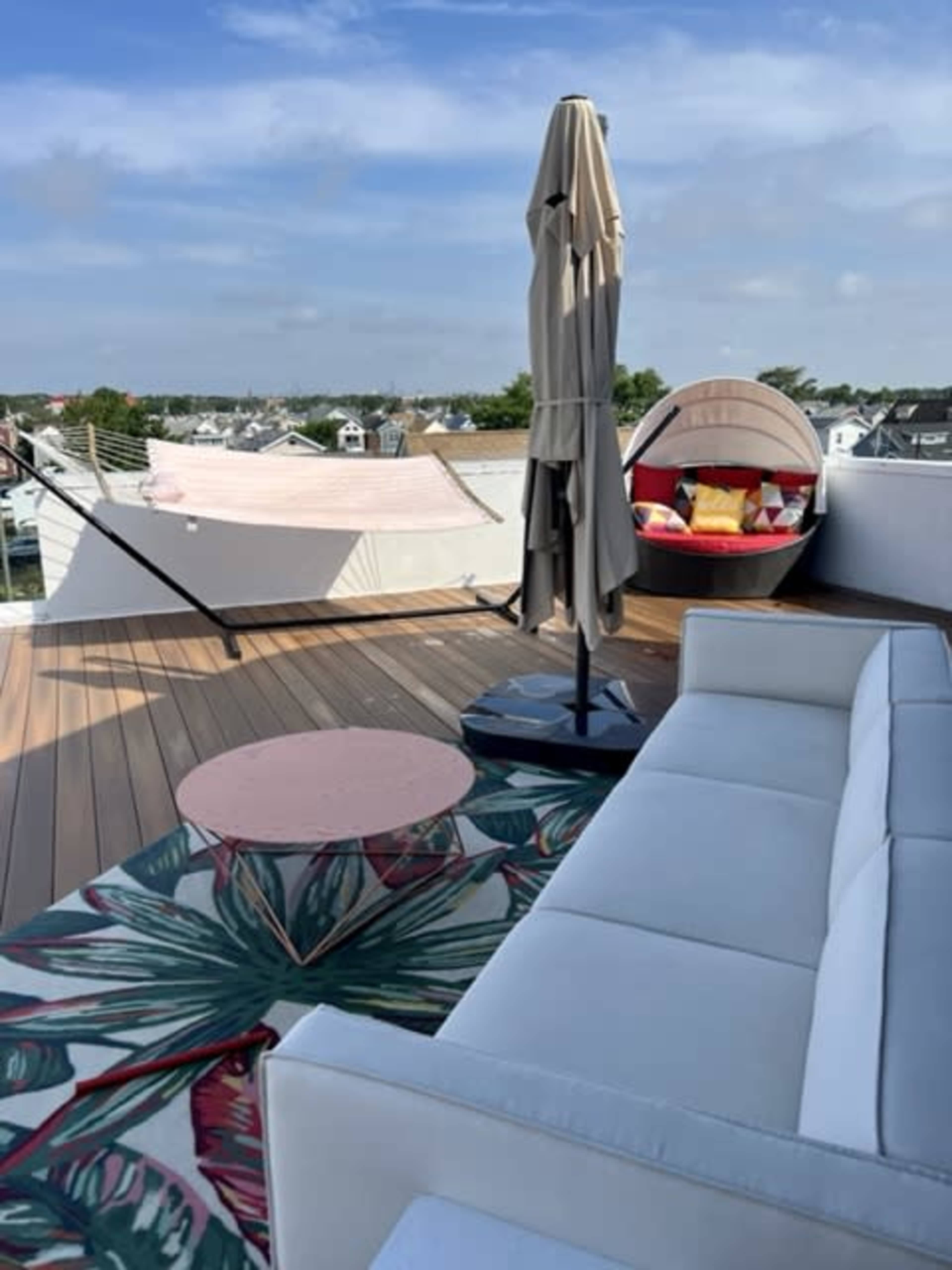 The image shows a rooftop deck featuring a white sofa, a pink circular table, a patio umbrella, a hanging chair, and a hammock, with a view of houses in the distance under a partly cloudy sky.