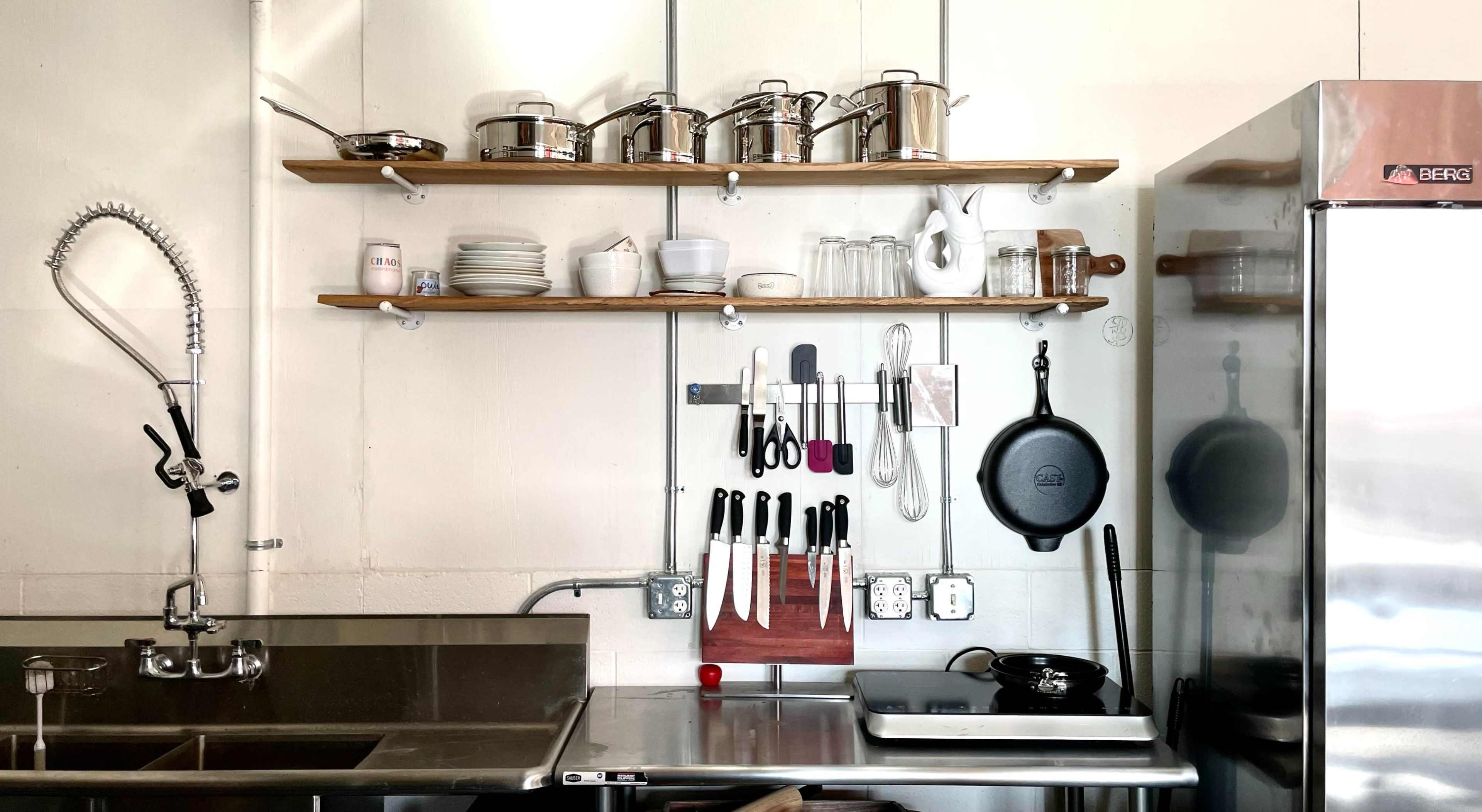 A kitchen with stainless steel countertops, shelves holding kitchenware, and a variety of utensils and knives neatly arranged.