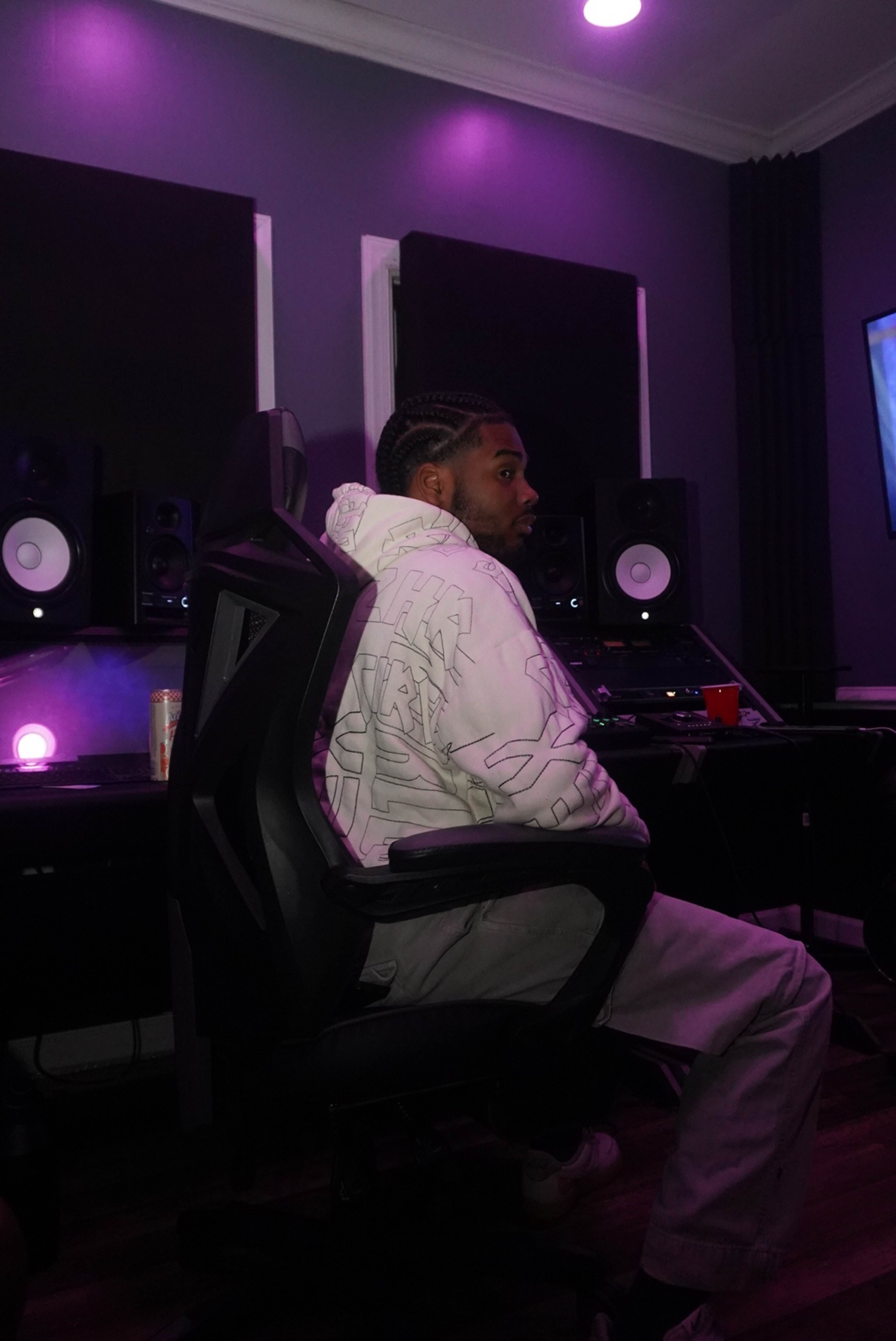 A person sitting in a chair in a music studio surrounded by speakers and dim purple lighting.
