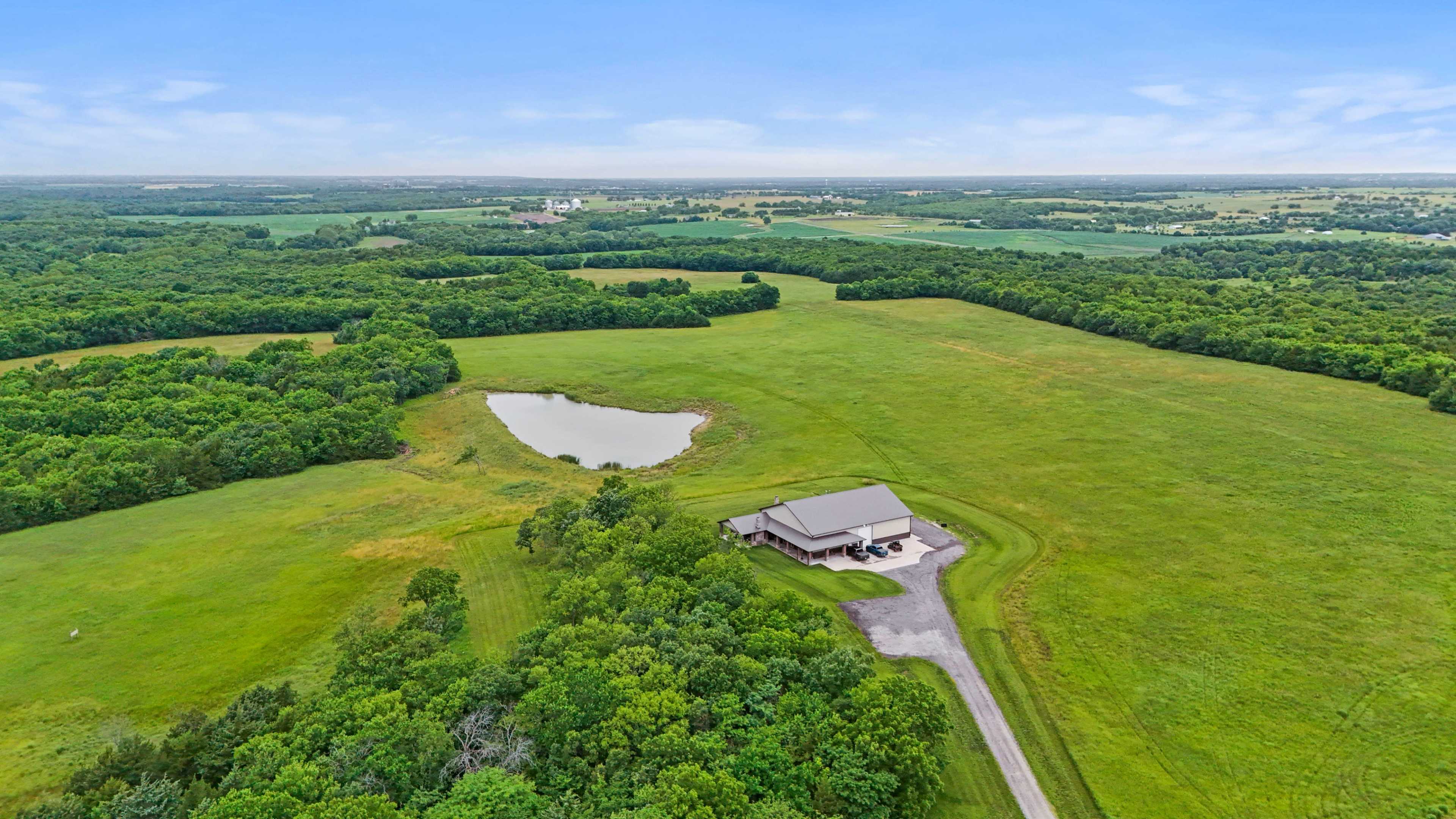 Aerial view of a large, green landscape featuring a house near a small pond surrounded by trees.