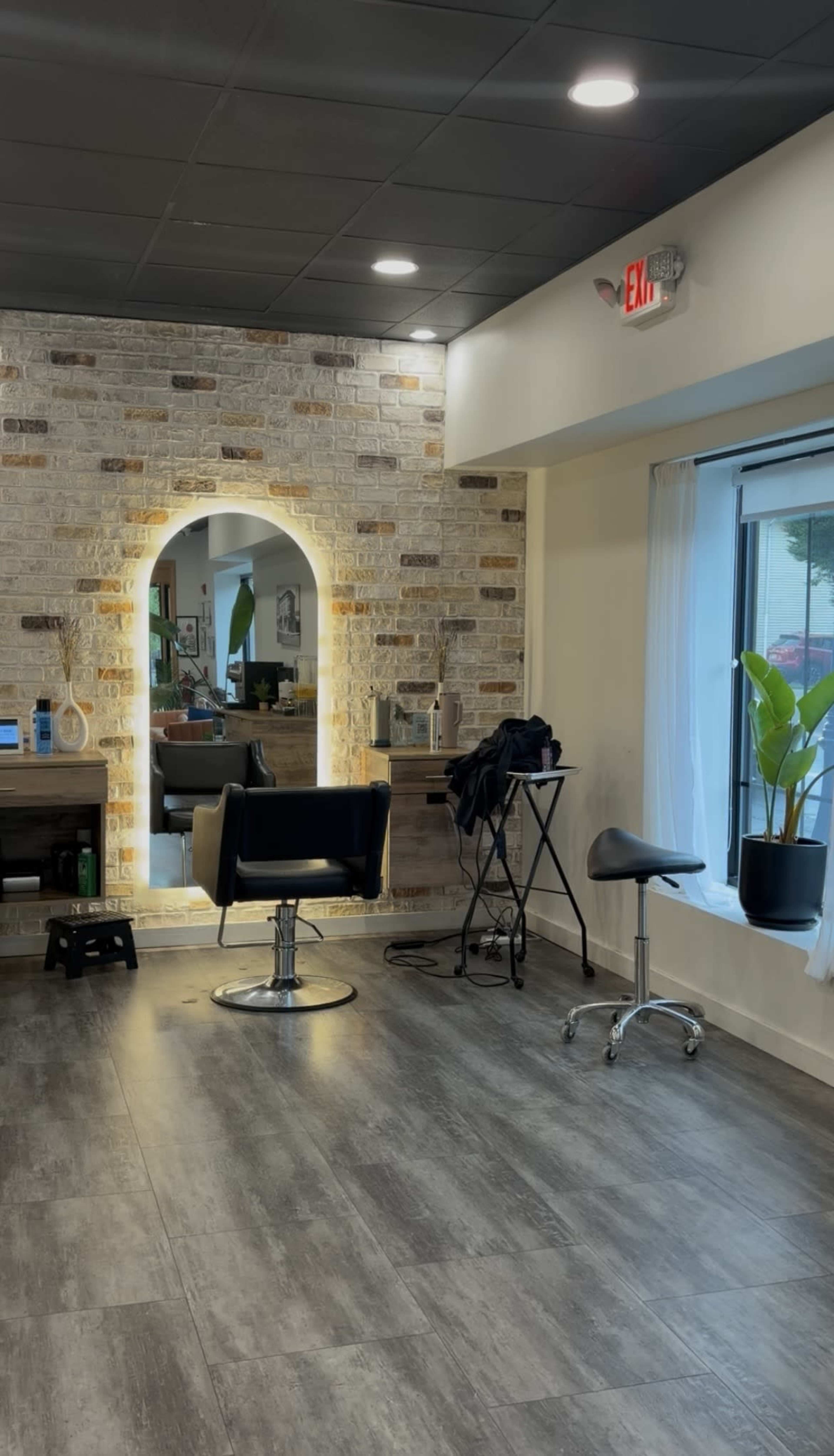The image shows a brightly lit hair salon featuring a brick wall, a styling chair, a mirror, and a potted plant by a window.