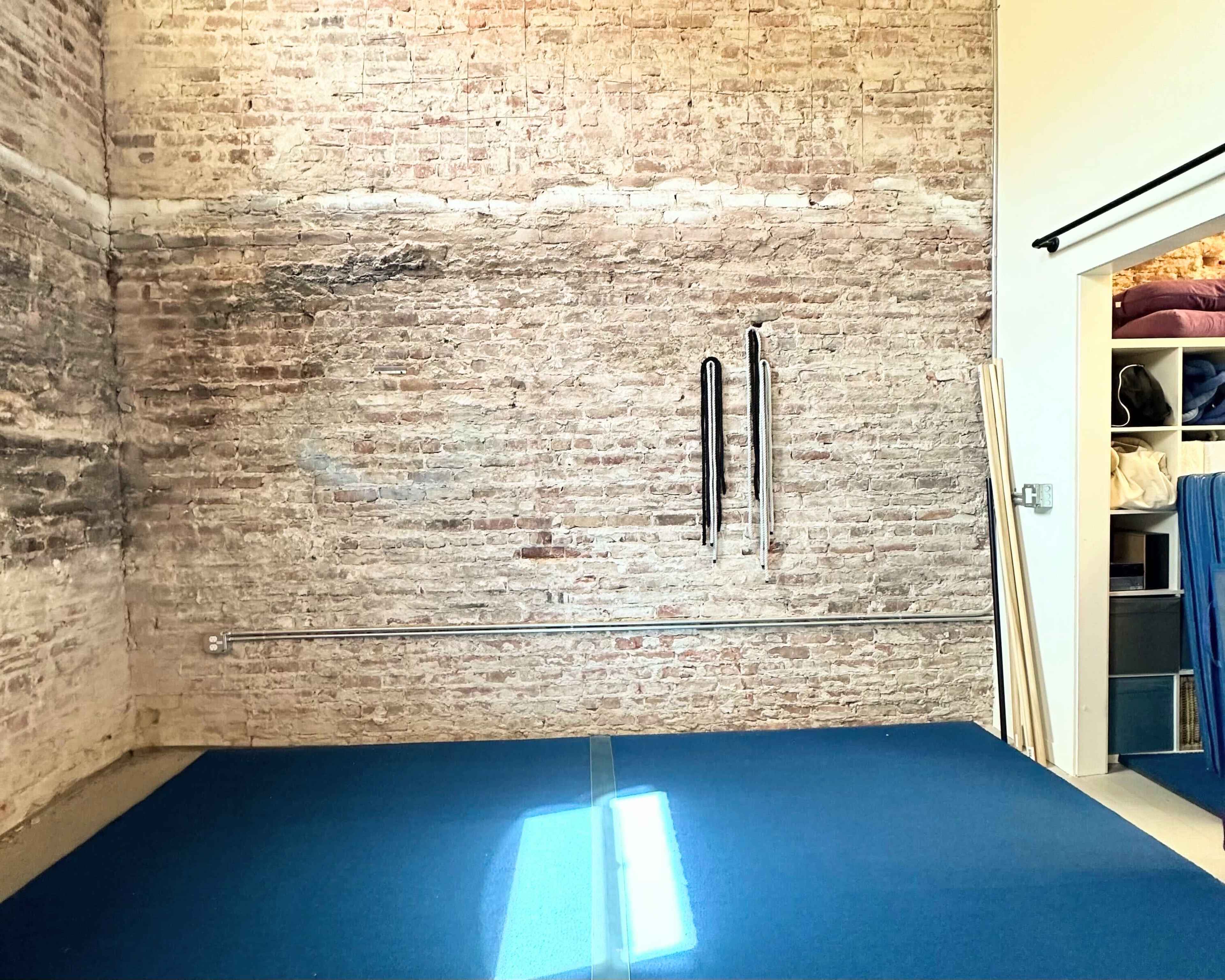 A room with a brick wall, a blue mat on the floor, and storage shelves filled with blankets and other items.