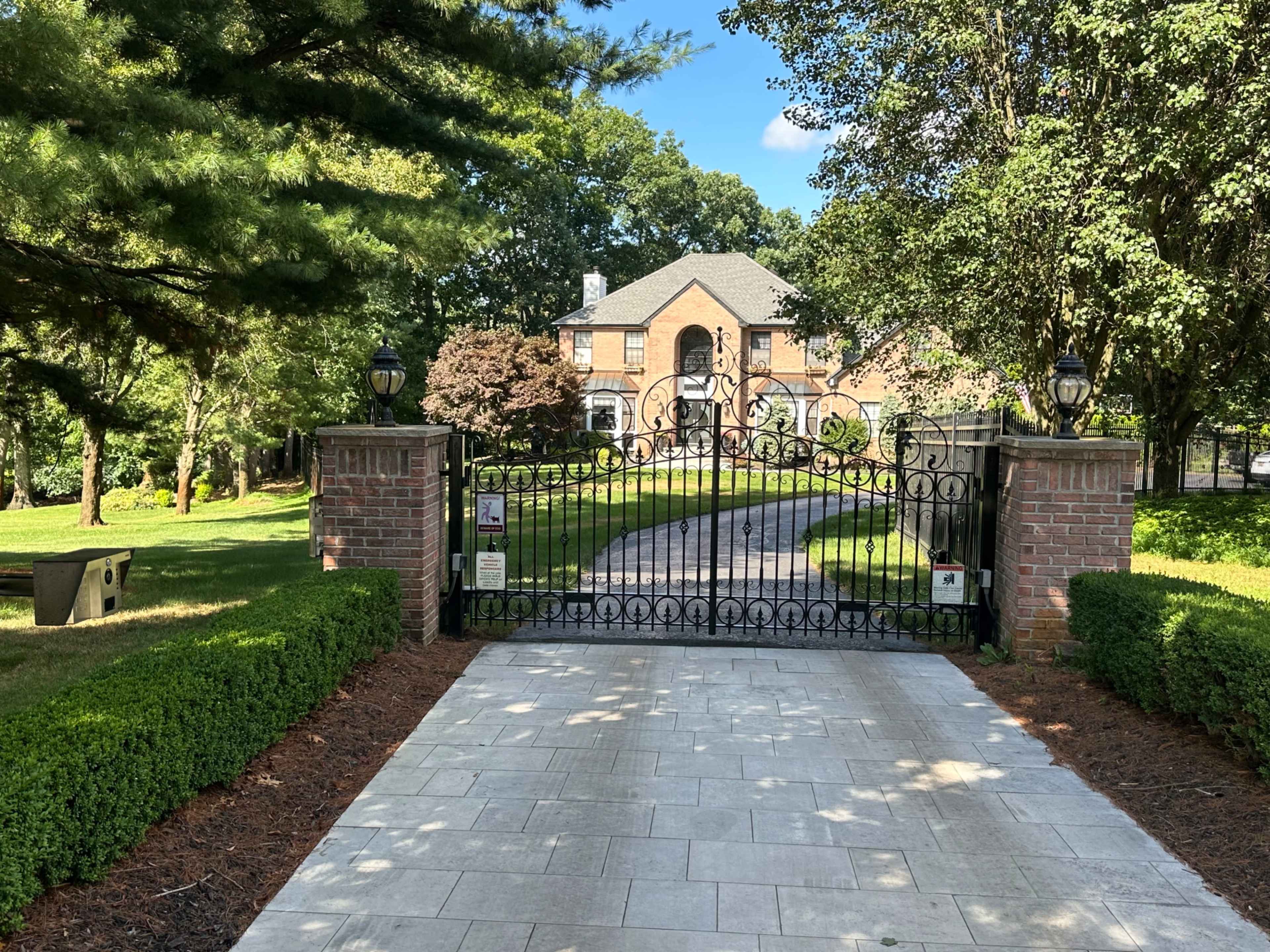 A wrought iron gate leads to a large brick house surrounded by greenery and trees.