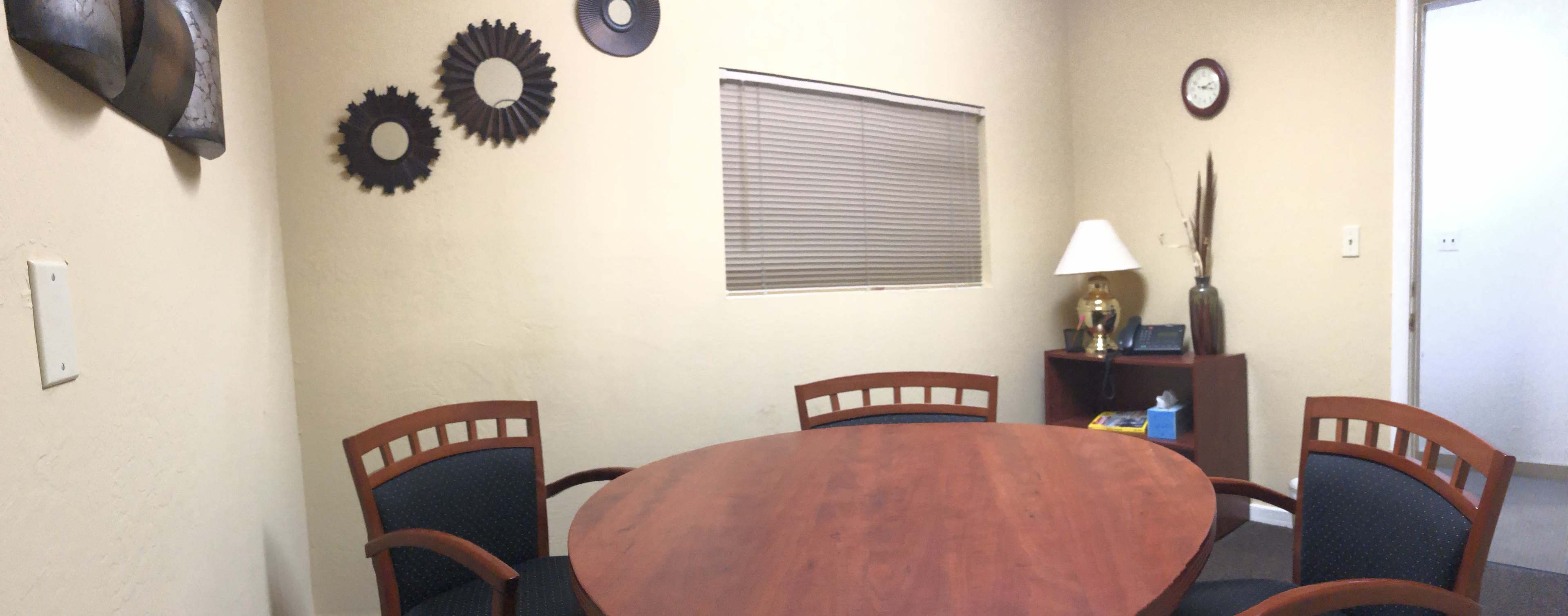 A small meeting room with a wooden table surrounded by three chairs, a lamp, and a clock on the wall.