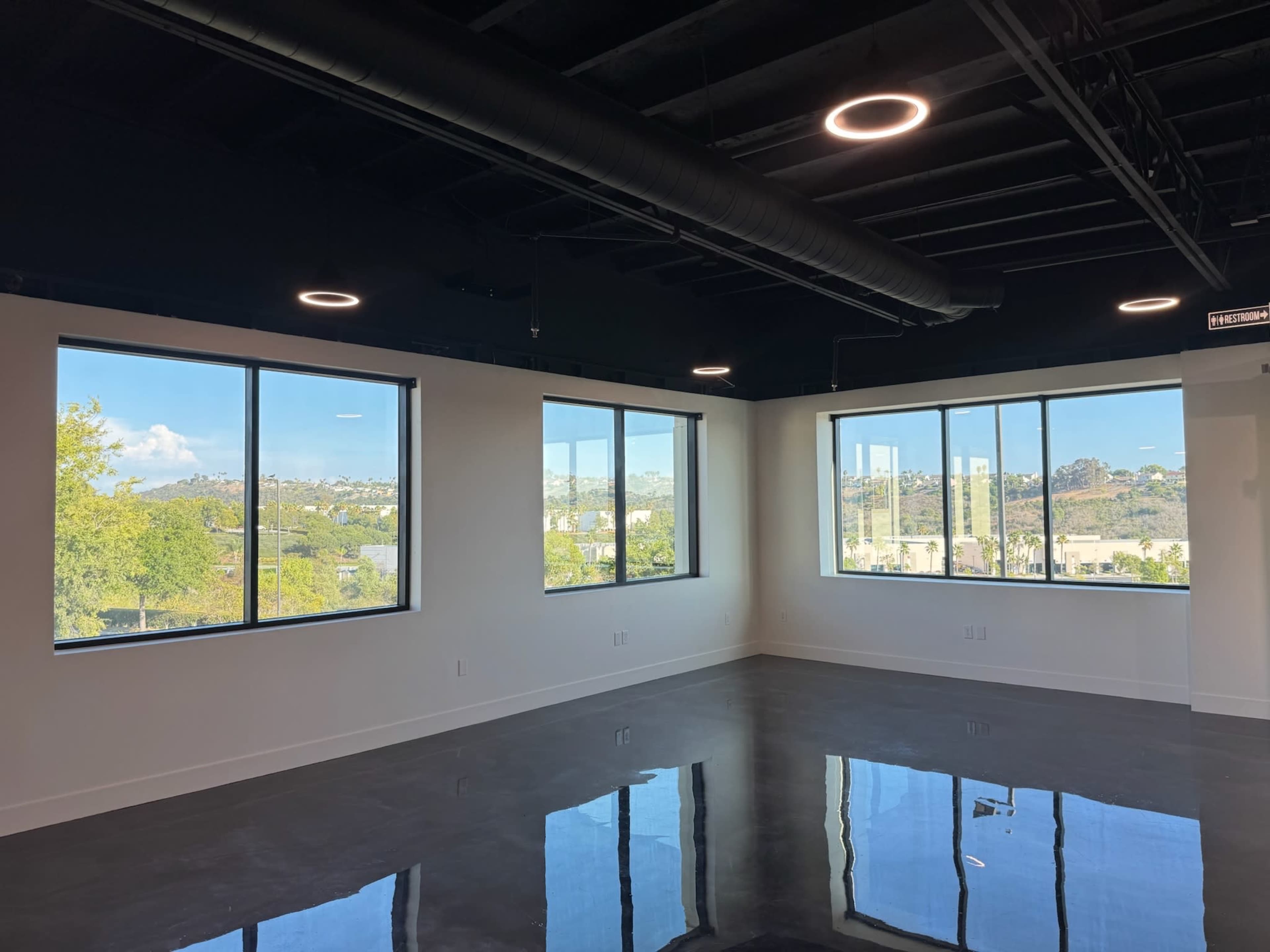The image shows a modern, empty office space with large windows that provide a view of an outdoor landscape and reflect a clear ceiling.
