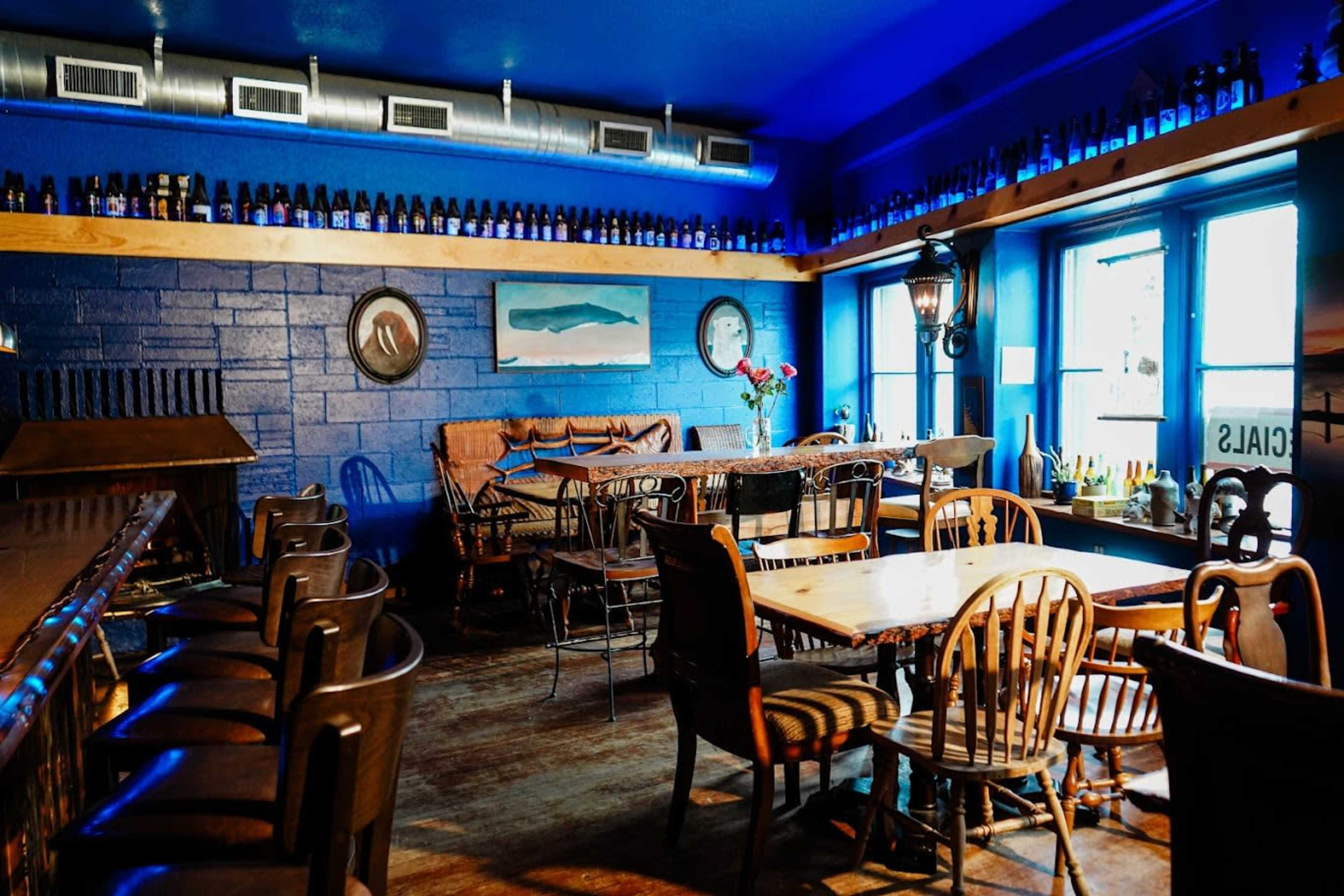 A cozy restaurant interior featuring blue walls, wooden tables, and various bottles displayed on a shelf above the windows.