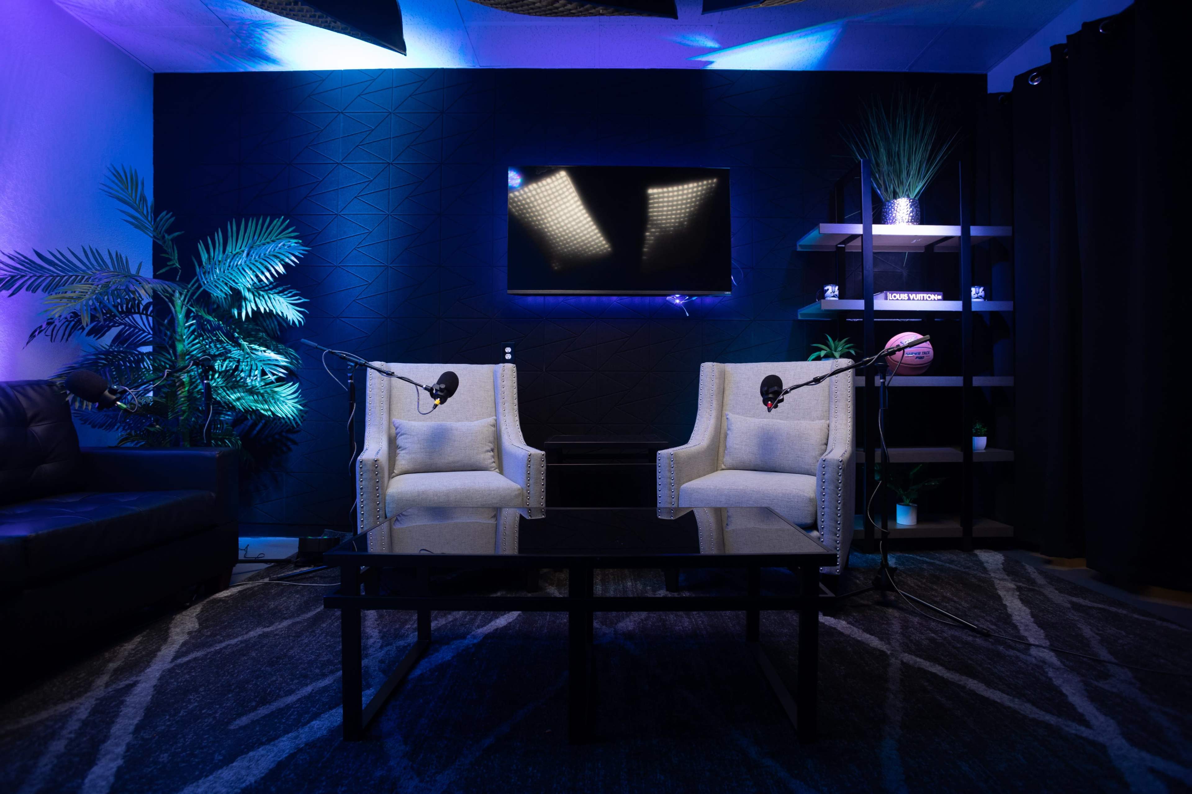 A modern podcast studio featuring two armchairs, a coffee table, microphones on stands, a large screen, and decorative plants under blue lighting.