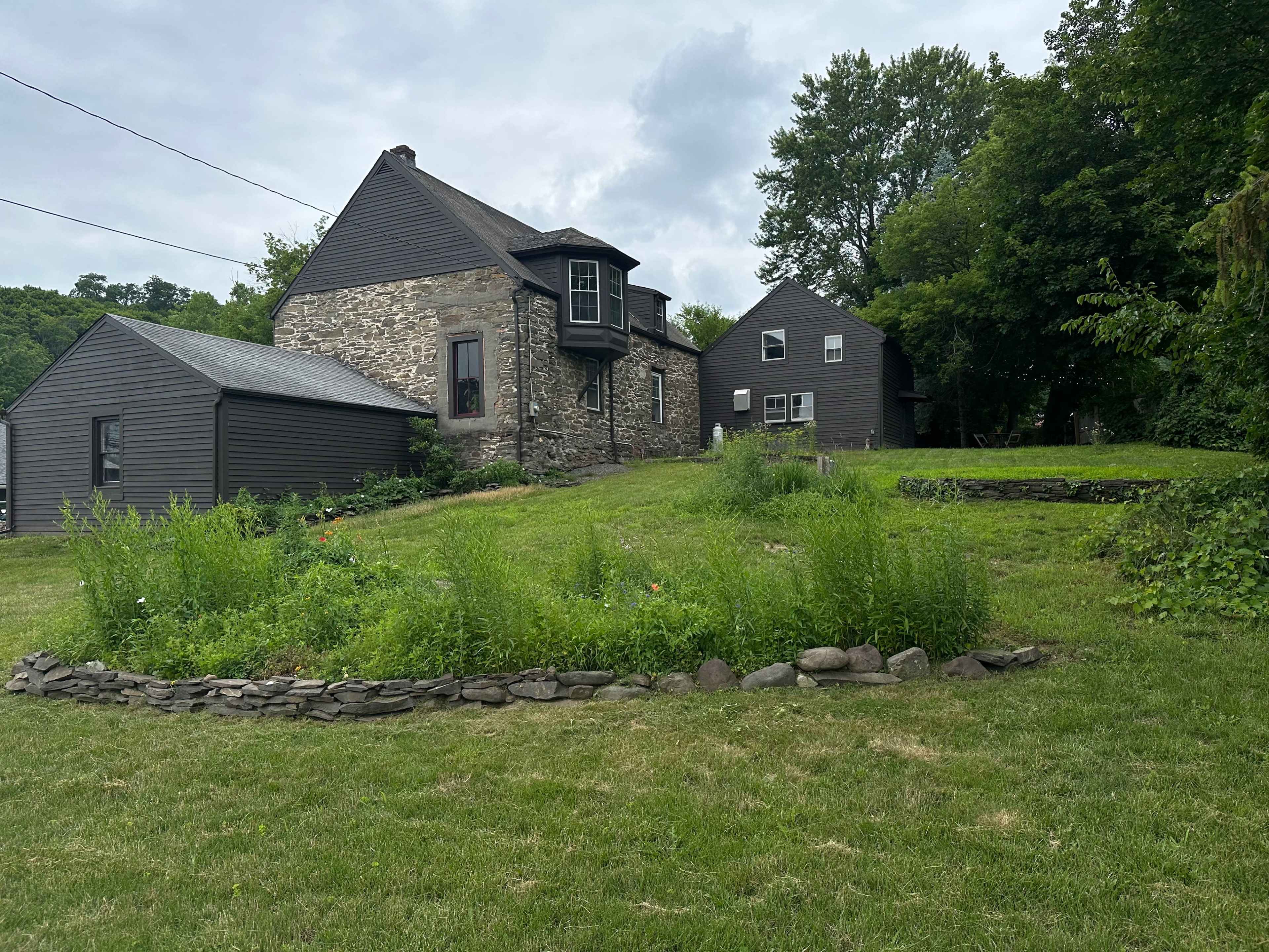 A grassy area featuring two houses, one made of stone and the other clad in dark siding, surrounded by trees and a stone-bordered garden.