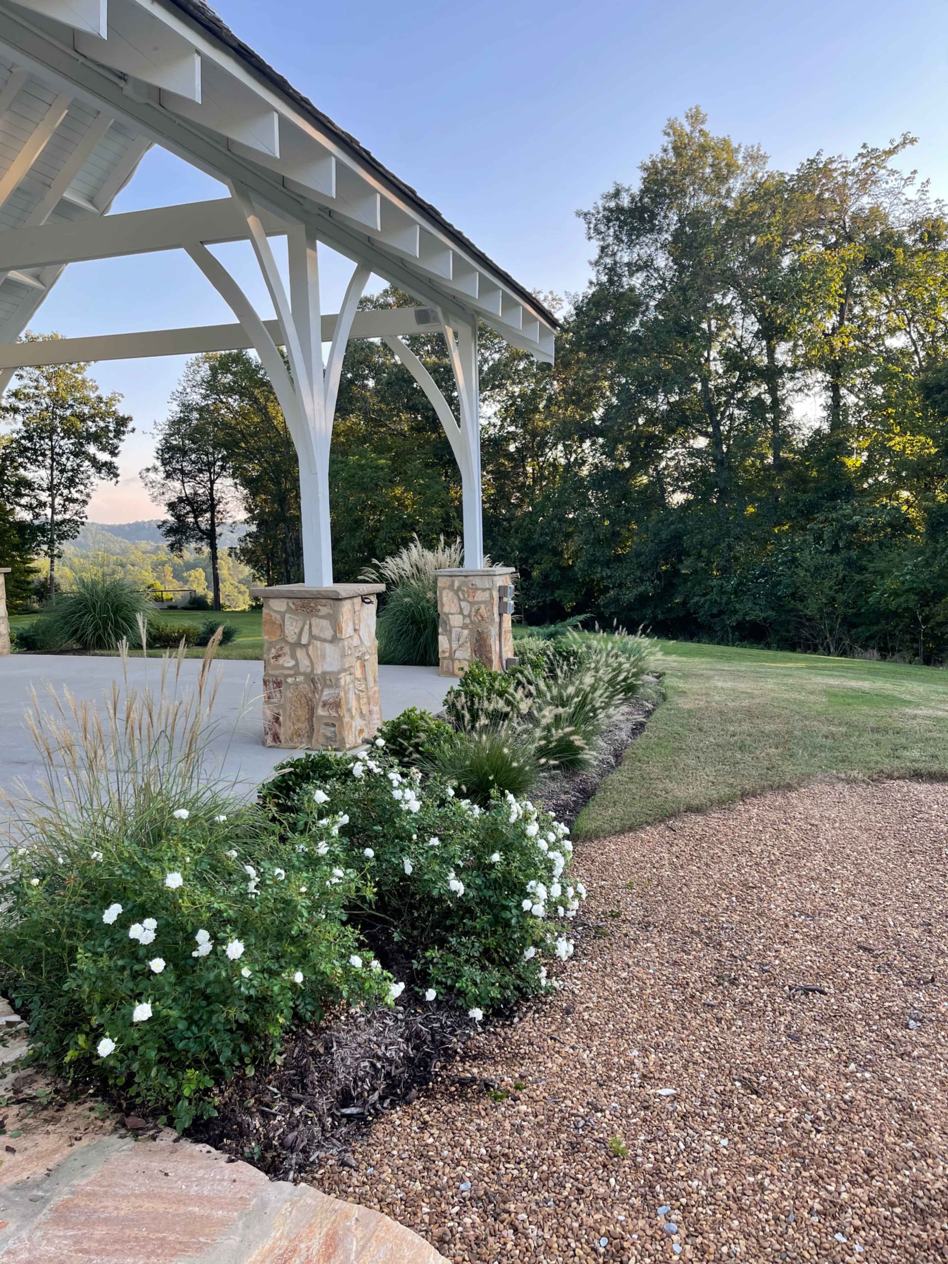 A covered patio area with stone pillars surrounded by landscaped greenery and white flowers, set against a backdrop of trees and a clear sky.
