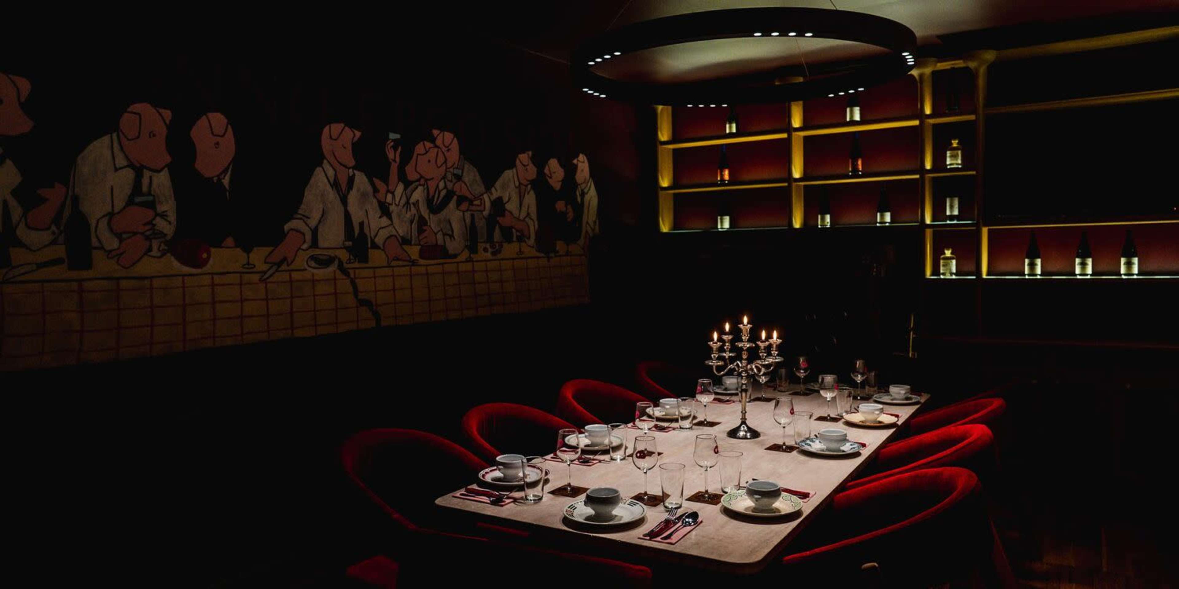 A long dining table is set with dishes and glassware in a dimly lit restaurant featuring artwork on the walls.